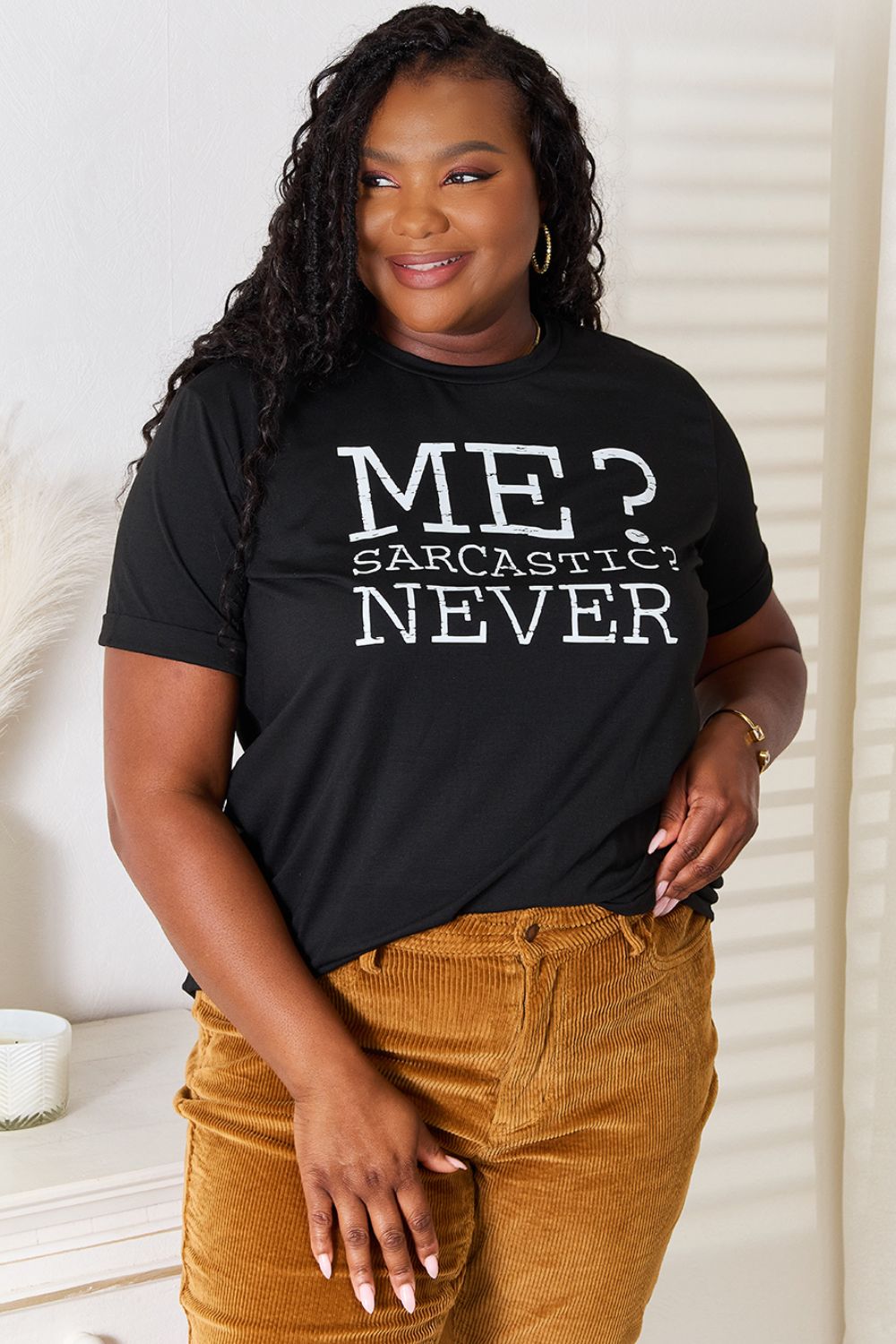 A woman with long dark hair smiles while wearing a trendy black Simply Love Letter Graphic Round Neck T-Shirt featuring the text "ME? Sarcastic? NEVER" and brown corduroy pants. She stands in a room with white walls and decor.