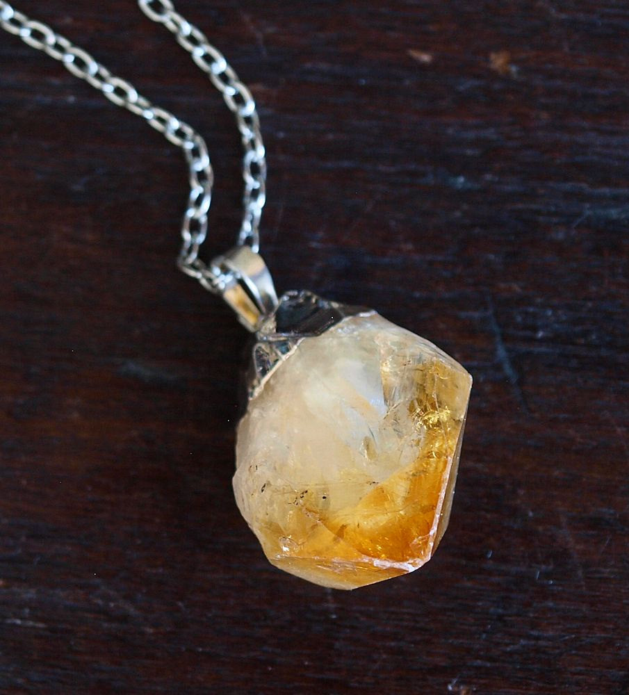 The Raw Citrine Crystal Statement Necklace features a rough citrine crystal and a silver-capped pendant on a shimmering silver chain, elegantly displayed against a black background.