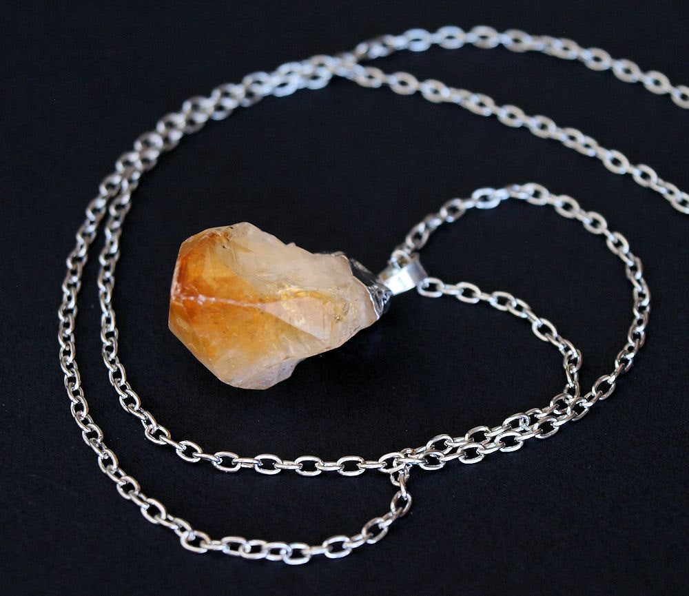 The Raw Citrine Crystal Statement Necklace features a rough citrine crystal and a silver-capped pendant on a shimmering silver chain, elegantly displayed against a black background.