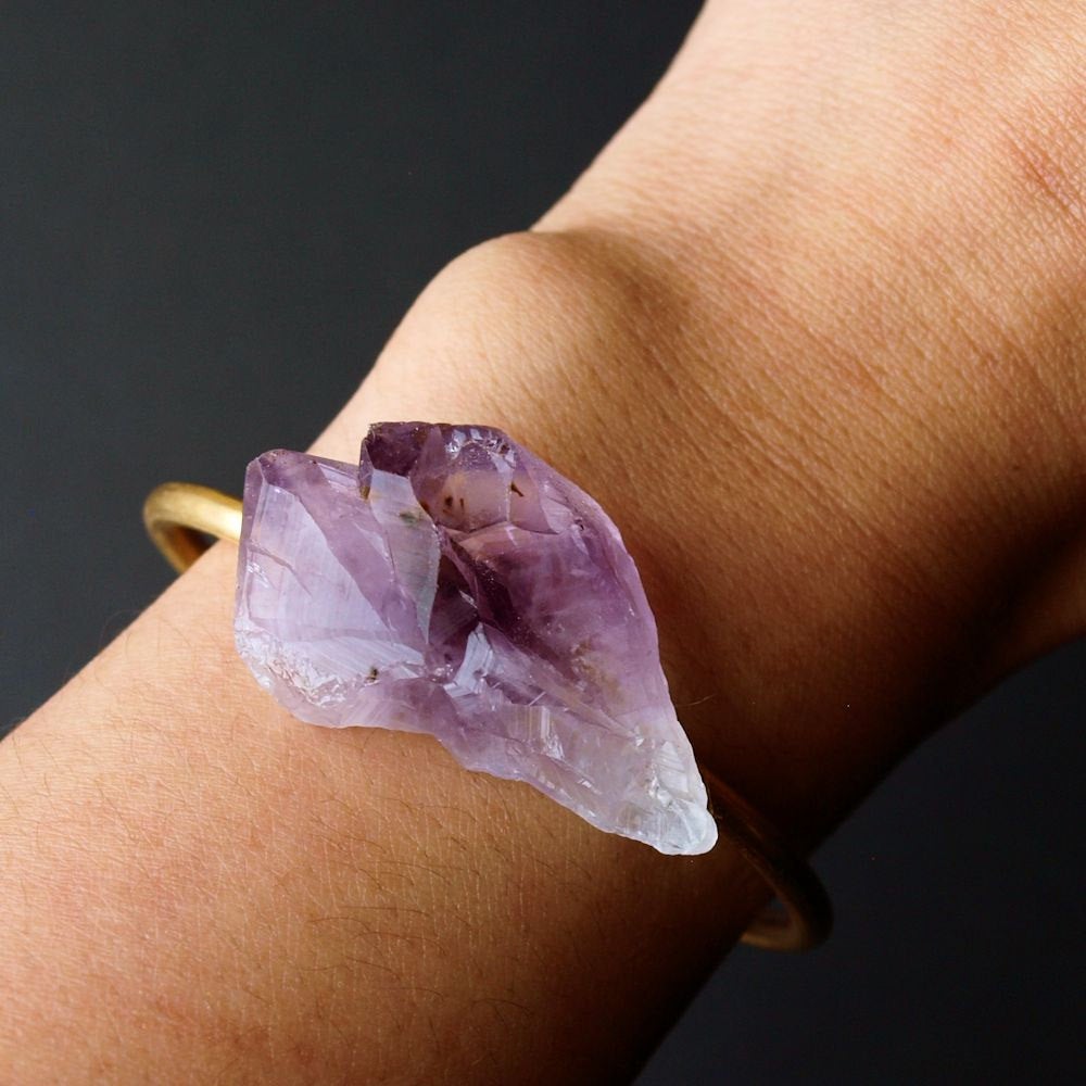 Close-up of a person's wrist wearing the Raw Amethyst Bangle, an adjustable bracelet with a large, rough purple stone set on a gleaming gold band.