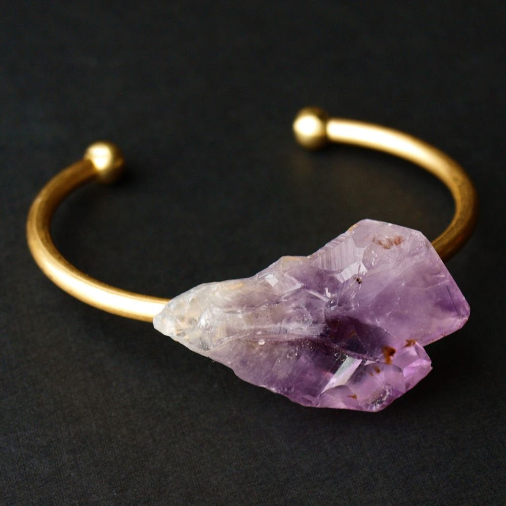 Close-up of a person's wrist wearing the Raw Amethyst Bangle, an adjustable bracelet with a large, rough purple stone set on a gleaming gold band.