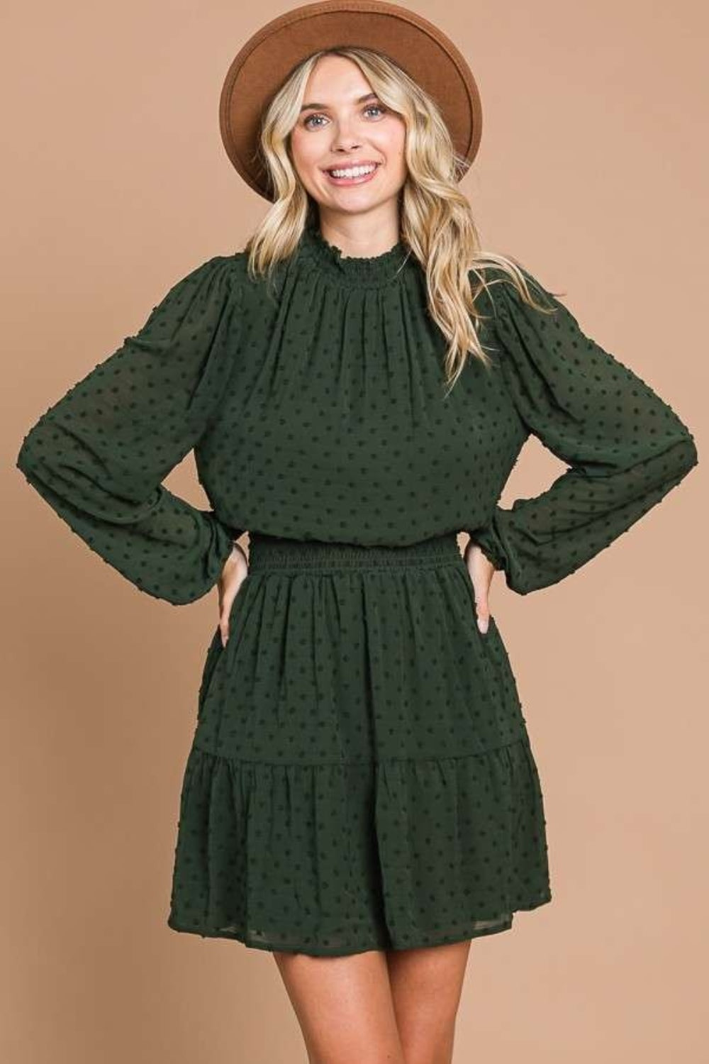 A woman wearing the Culture Code Swiss Dot Smock Neck Mini Dress with Pockets in dark green, featuring long sleeves, is standing with her back to the camera.