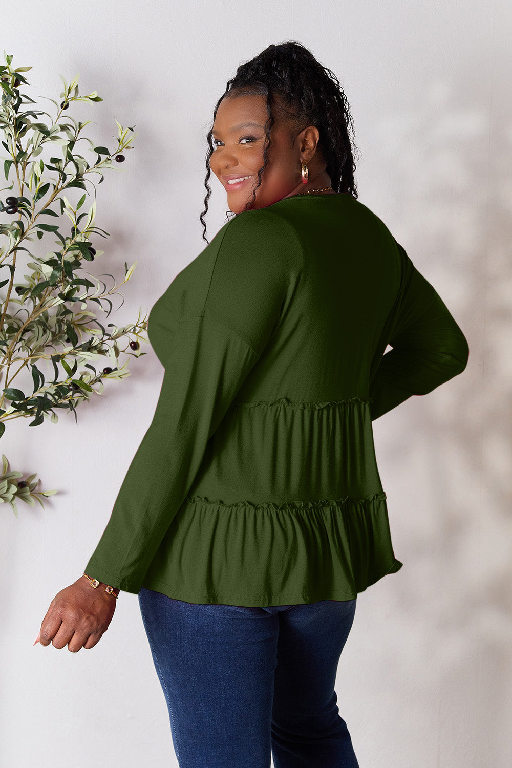 Person stands indoors near a plant, wearing the Double Take Half Button Long Sleeve Ruffle Hem Blouse in gray and blue jeans, smiling while looking to the side. The slightly stretchy fabric of the blouse adds comfort while its buttoned design offers a touch of elegance.