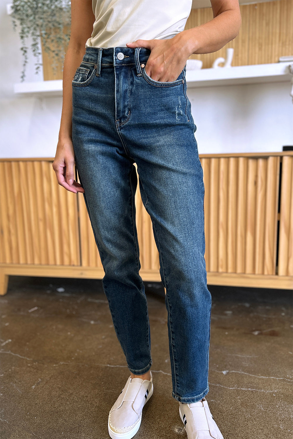 Person wearing Judy Blue Full Size Tummy Control High Waist Slim Jeans in blue and a white shirt, with their hands in their pockets. They are standing indoors on a polished concrete floor with a wooden cabinet and plant in the background.