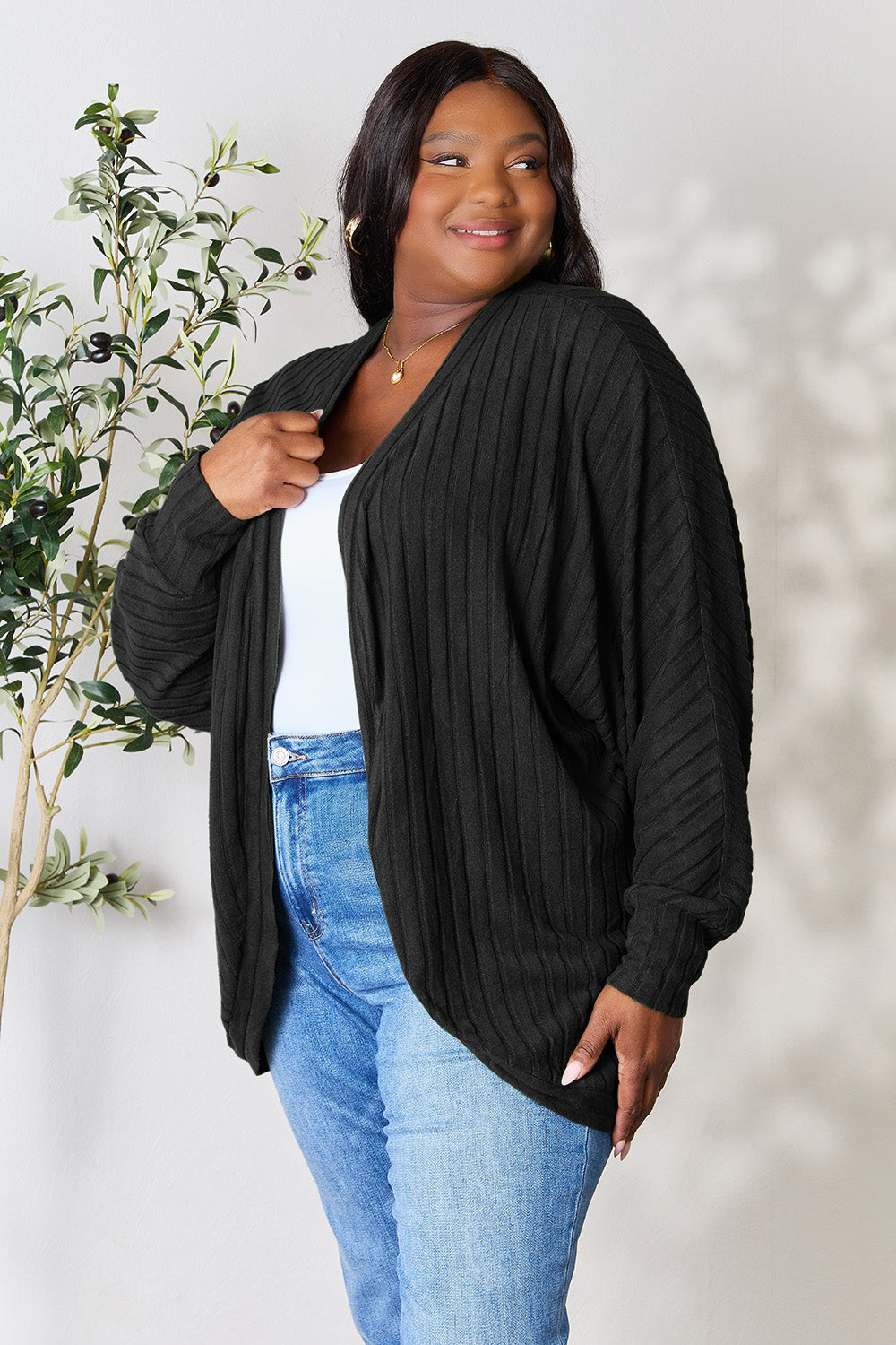 A person with wavy hair, wearing the Basic Bae Full Size Ribbed Cocoon Cardigan over a white top and beige pants, stands in front of a light-colored background with a leafy plant, showcasing an effortlessly basic style.