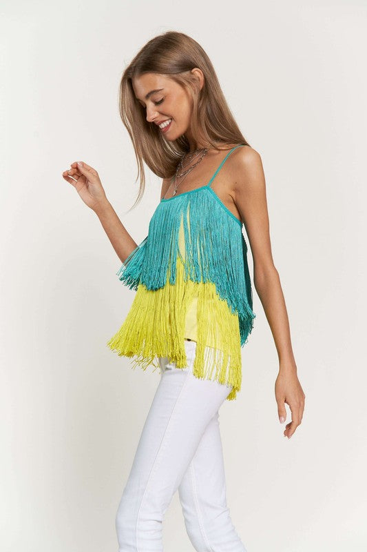 A woman with long brown hair is wearing a Fringe Overlay Cross Straps Party Cami Top featuring blue and yellow layers, paired with white pants, standing against a plain background.