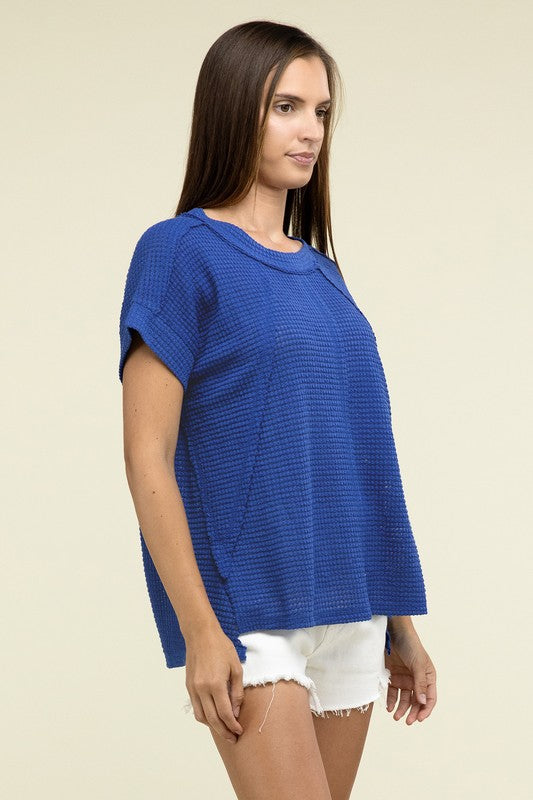 A woman with long brown hair is wearing a purple Brushed Waffle Exposed-Seam Short Sleeve Top and blue jeans, standing against a plain beige background. This casual wardrobe choice, with its short sleeves and side slits, is perfect for everyday wear.