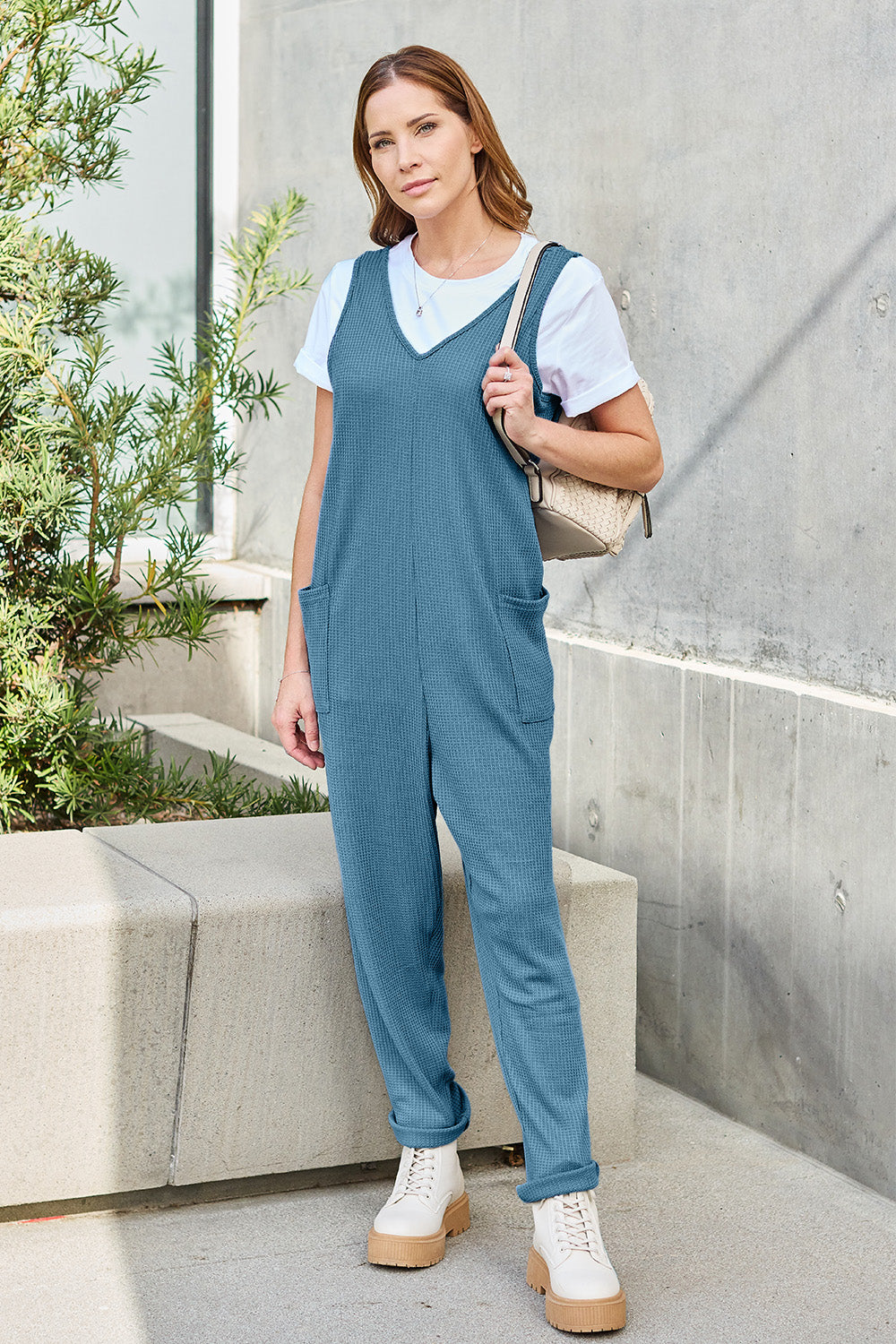 A person stands outdoors wearing the Double Take Full Size Sleeveless Straight Jumpsuit in blue and brown sandals, holding a cream-colored bag. In the background, there's a wooden bench and palm trees.