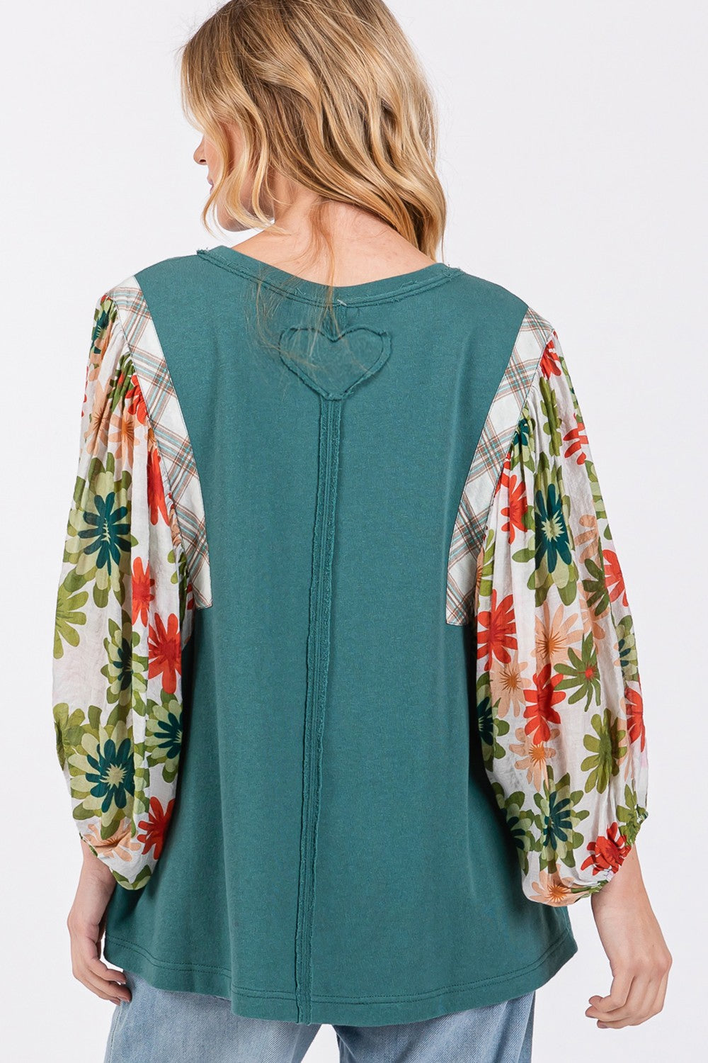 A person in a hat wears the SAGE + FIG Full Size Printed Balloon Sleeve Contrast Top with teal floral-patterned sleeves, blue jeans, and a back heart patch detail.