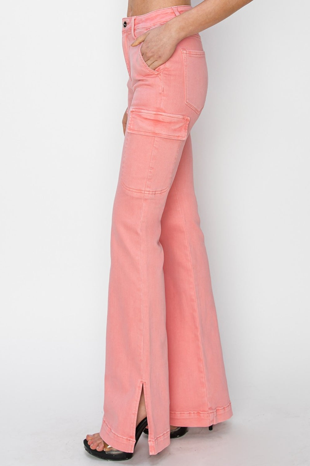 A person wearing RISEN Full Size High Rise Side Slit Cargo Bootcut Jeans, featuring a pink flared design with front pockets, stands against a plain background.