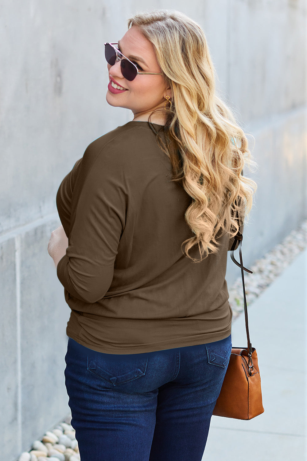 A woman in a slightly stretchy, brown Basic Bae Full Size Round Neck Batwing Sleeve top and blue jeans stands against a concrete wall, holding a brown handbag.