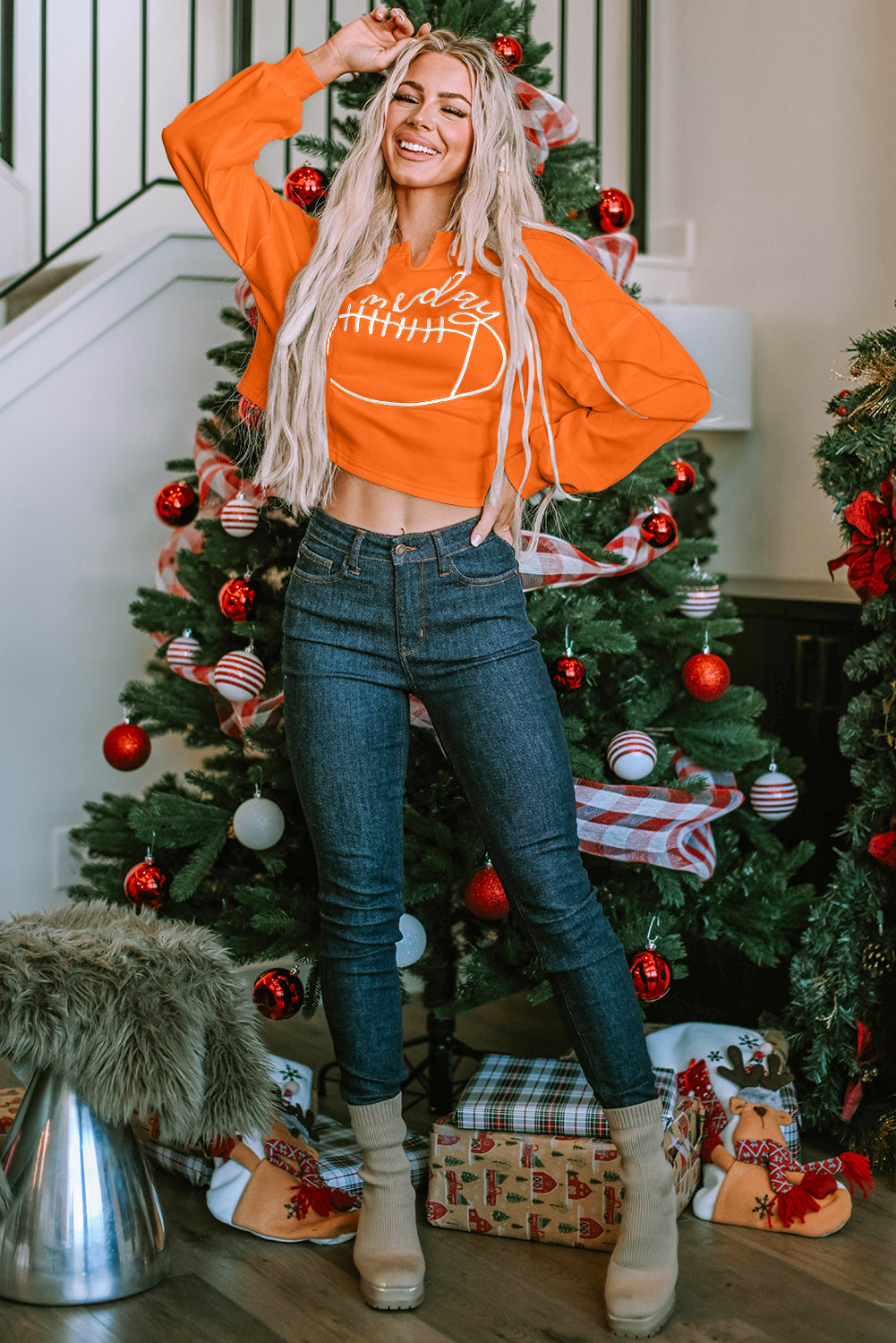 A woman with blonde hair, wearing an Orange Game Day Lettering Rugby Football Notched Neck Sweatshirt and blue jeans, stands next to a decorated Christmas tree.