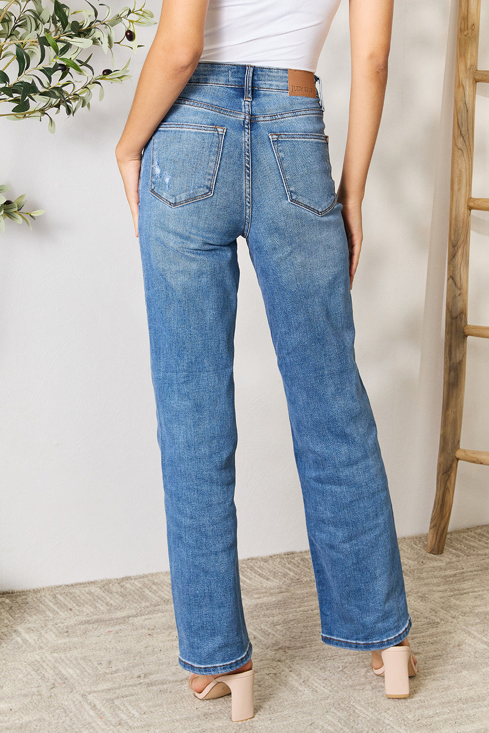 A person wearing a white top and Judy Blue Full Size High Waist Distressed Jeans stands in a room with a ladder and a plant.