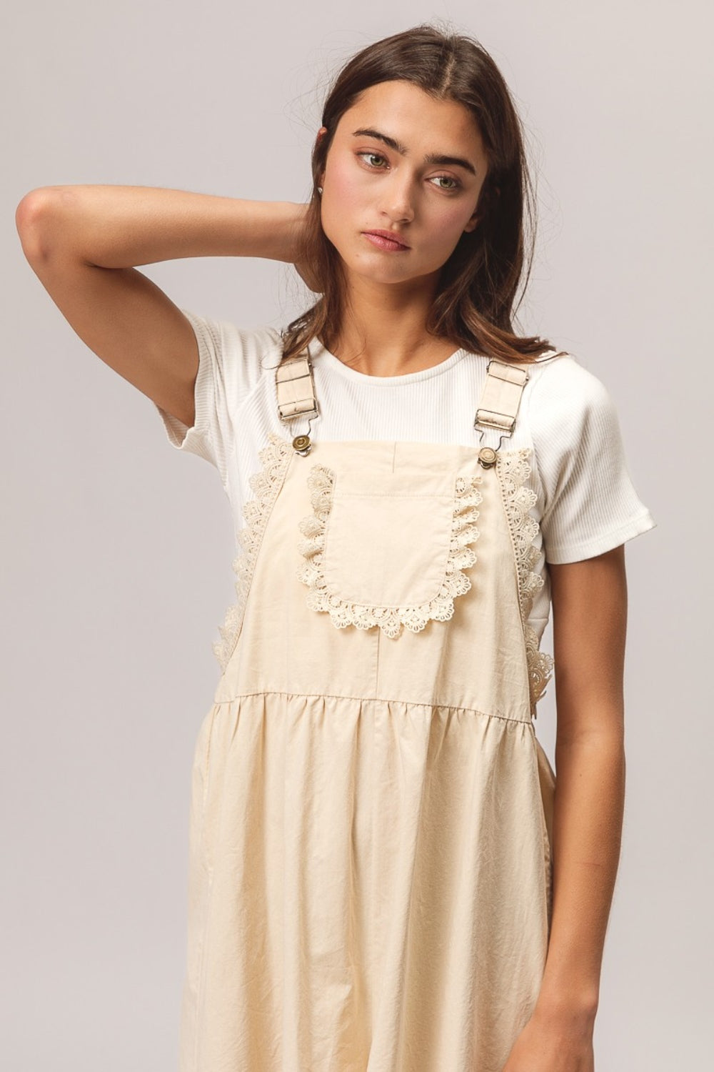 A young woman with long dark hair is sitting on a wooden stool, wearing a white t-shirt paired with the BiBi Back Patch Pocket Lace Trim Rompers—a cream-colored overall dress featuring back patch pockets. With a neutral expression set against a plain light gray background, she effortlessly embodies the perfect addition to any summer wardrobe collection.