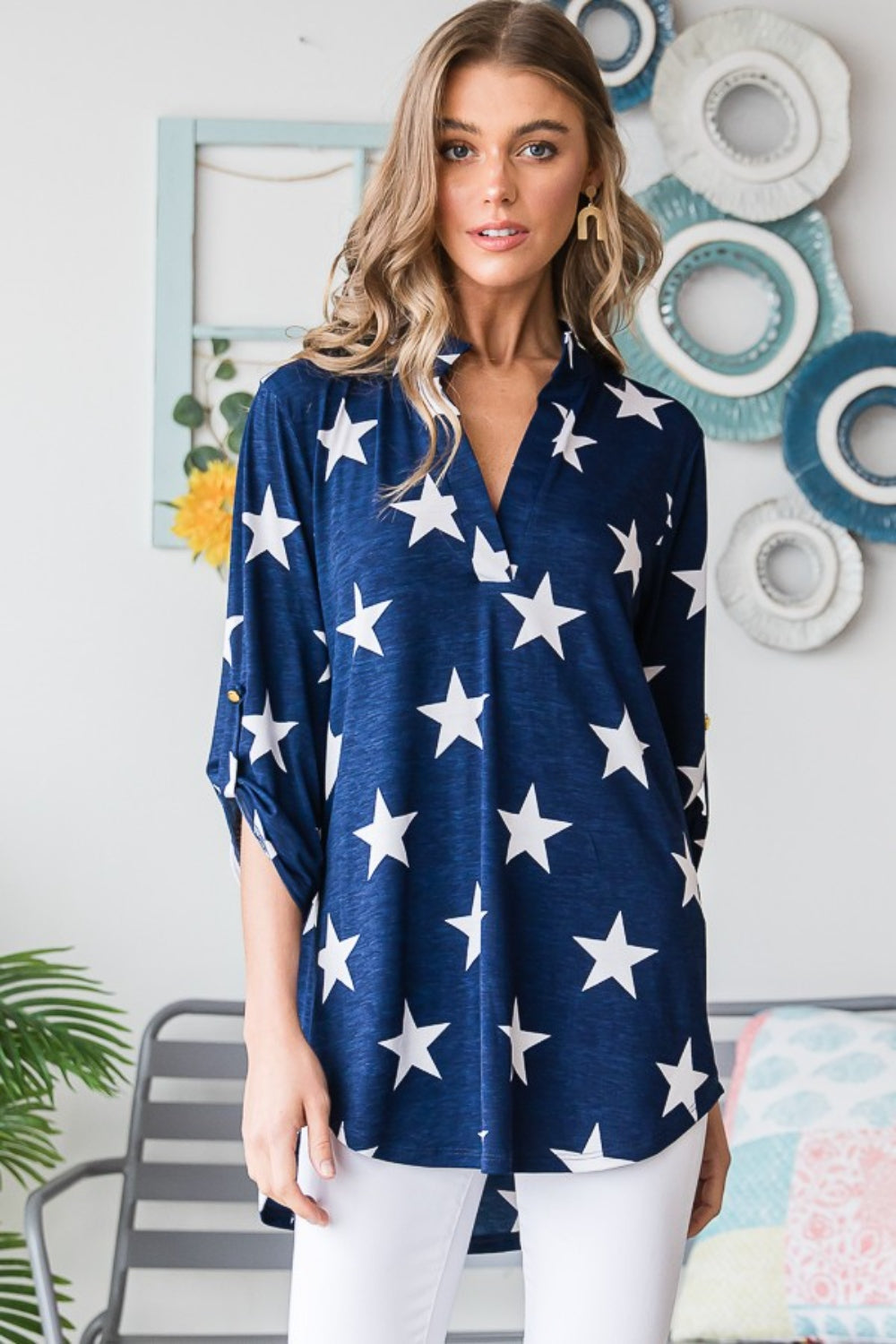 A woman with long hair is sporting the Heimish Full Size Roll-Tab Sleeve Star Print Top in navy blue, paired with white pants, perfect for casual outings. She stands in front of a light-colored wall adorned with round hangings.