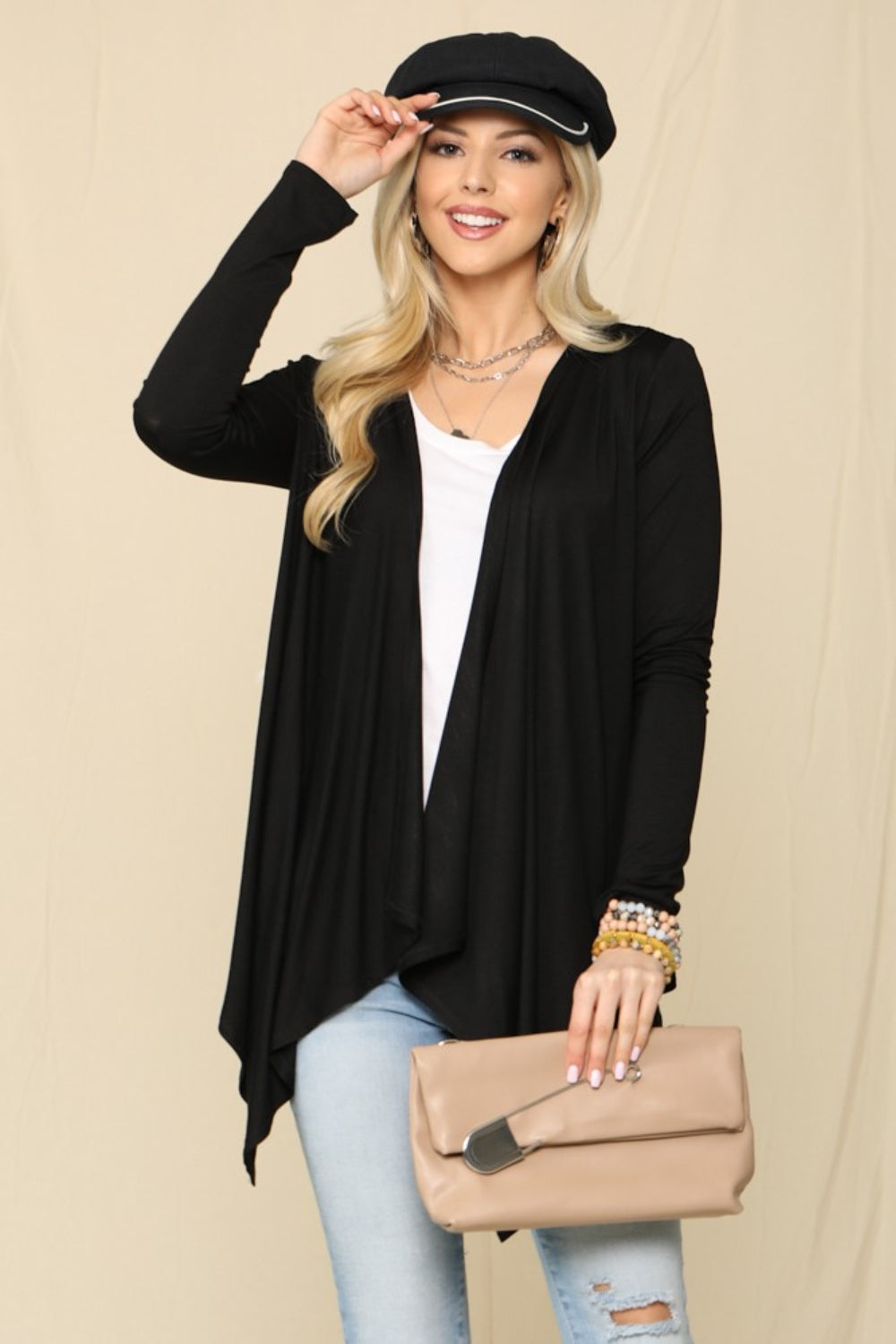 A woman wearing a black hat, the Celeste Full Size Open Front Knit Cardigan—an essential wardrobe staple—and a white top, holding a beige clutch, poses against a beige background.