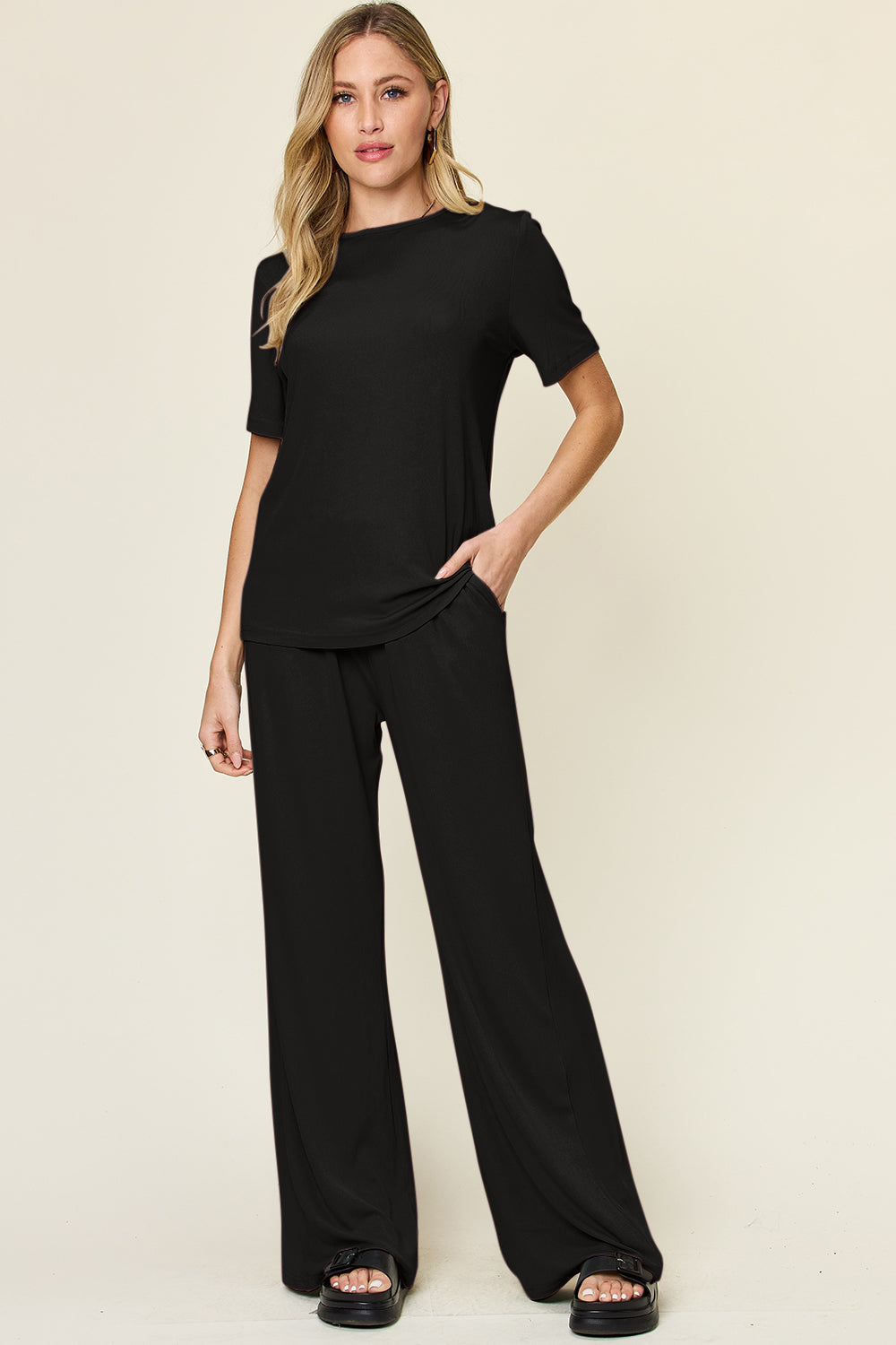 A person stands wearing the Double Take Full Size Round Neck Short Sleeve T-Shirt and Wide Leg Pants Set in a stretchy light grey fabric, with one hand in a pocket and black sandals.