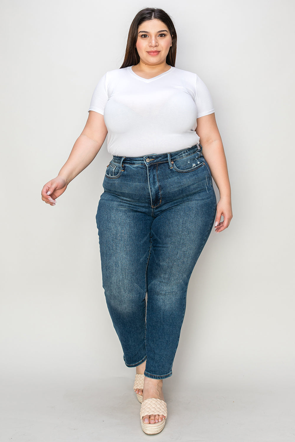 Person wearing Judy Blue Full Size Tummy Control High Waist Slim Jeans in blue and a white shirt, with their hands in their pockets. They are standing indoors on a polished concrete floor with a wooden cabinet and plant in the background.