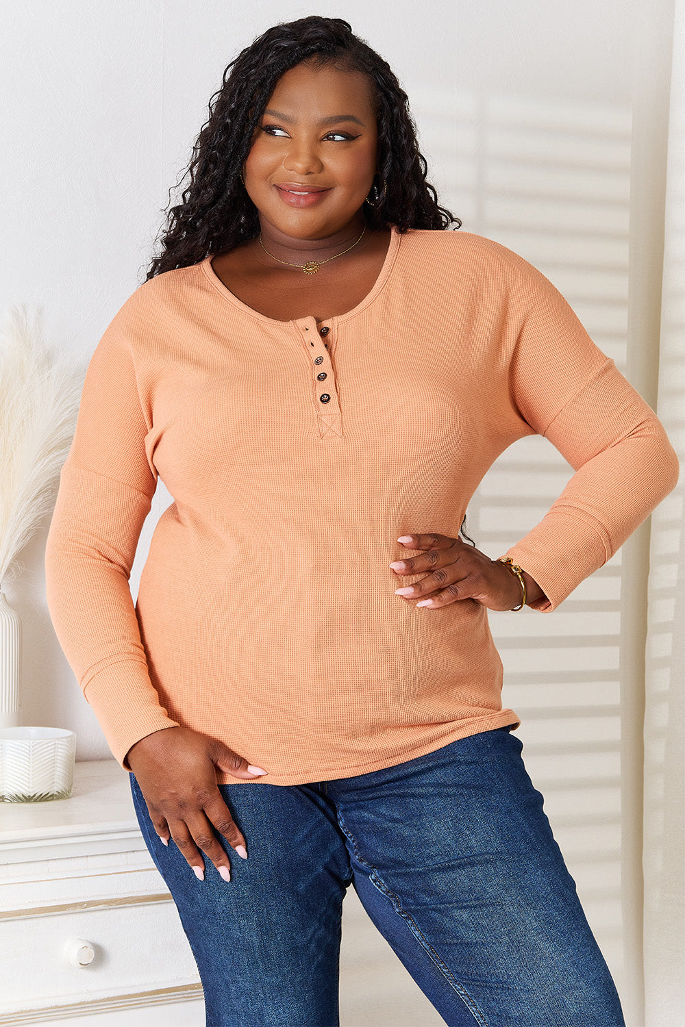 A woman stands indoors, wearing a Basic Bae Half Button Long Sleeve Top in peach and blue jeans, with her hand resting on her hip, showcasing versatile chic wardrobe choices.