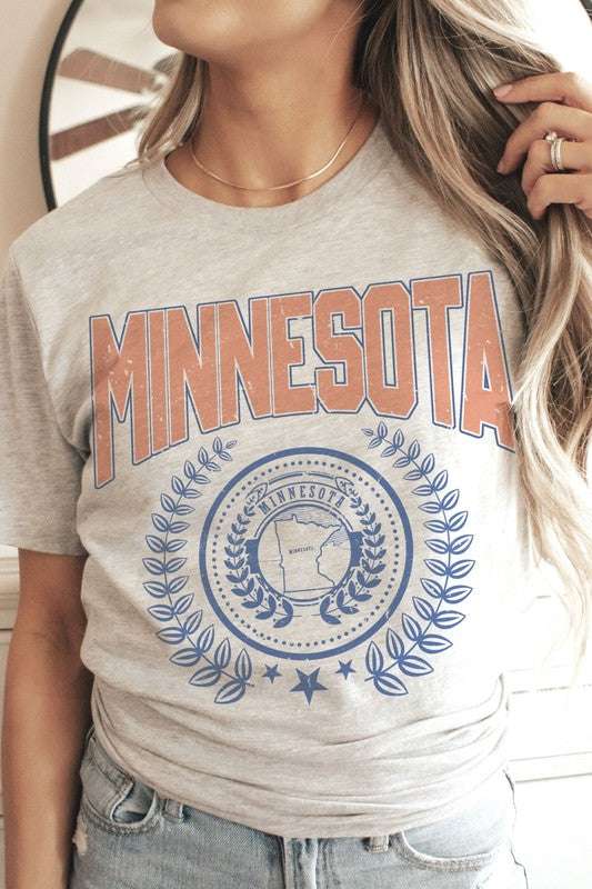 A woman sporting a 100% cotton MINNESOTA Graphic Tee with "Minnesota" emblazoned in bold orange letters and a decorative state emblem underneath, complemented by high-waisted denim shorts and a plaid shirt casually draped over her shoulders. The tee offers unisex sizing for an extra comfortable fit.