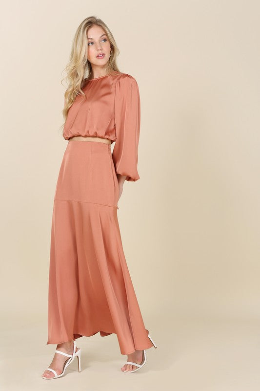 A woman stands against a plain background, elegantly adorned in the "Dressed up satin two-piece mermaid dress set" featuring a peach-colored, balloon-sleeve crop top and matching high-waisted maxi skirt, accessorized with white high-heeled sandals.