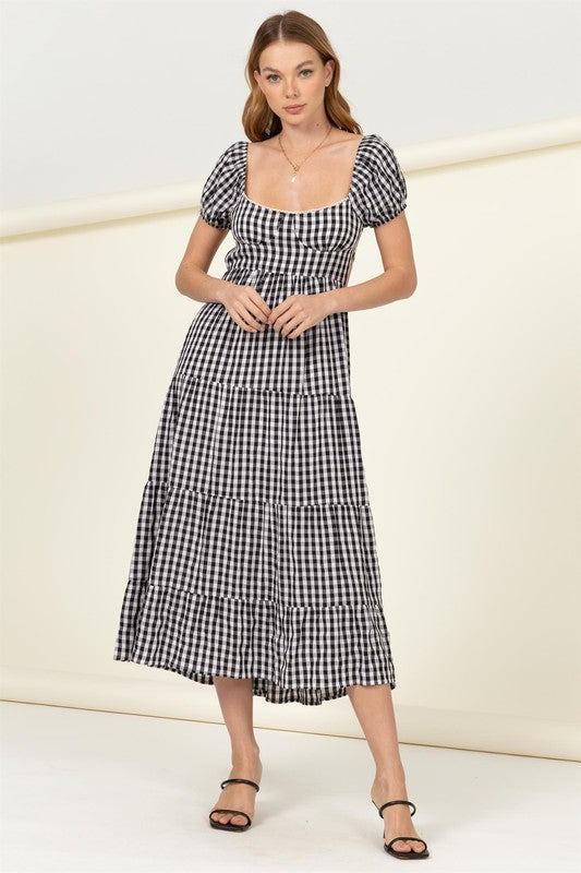 Wearing the "Somewhere to Go Tie-Back Gingham Print Maxi Dress," which features a black and white gingham print with puffed sleeves, a tiered skirt, and elegant tie-back detail, a person poses stylishly paired with black sandals.