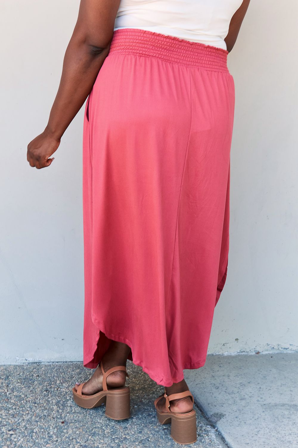 A person wearing a white top and the Doublju Comfort Princess Full Size High Waist Scoop Hem Maxi Skirt in an elegant coral color, paired with tan sandals, stands in front of a plain wall.