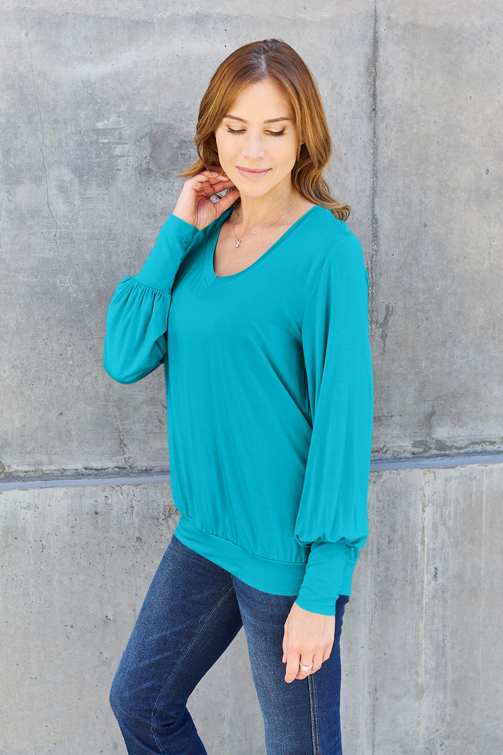 A woman with brown hair is standing against a concrete wall, wearing the Basic Bae Full Size V-Neck Lantern Sleeve Top in teal and blue basic-style jeans, smiling.