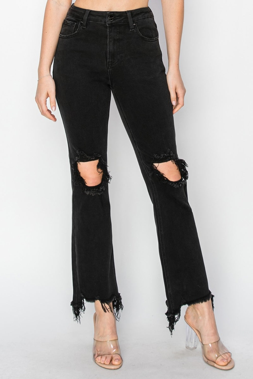 Rear view of a person wearing RISEN Full Size Distressed Raw Hem Jeans with Pockets in black, paired with clear-heeled sandals against a plain background.