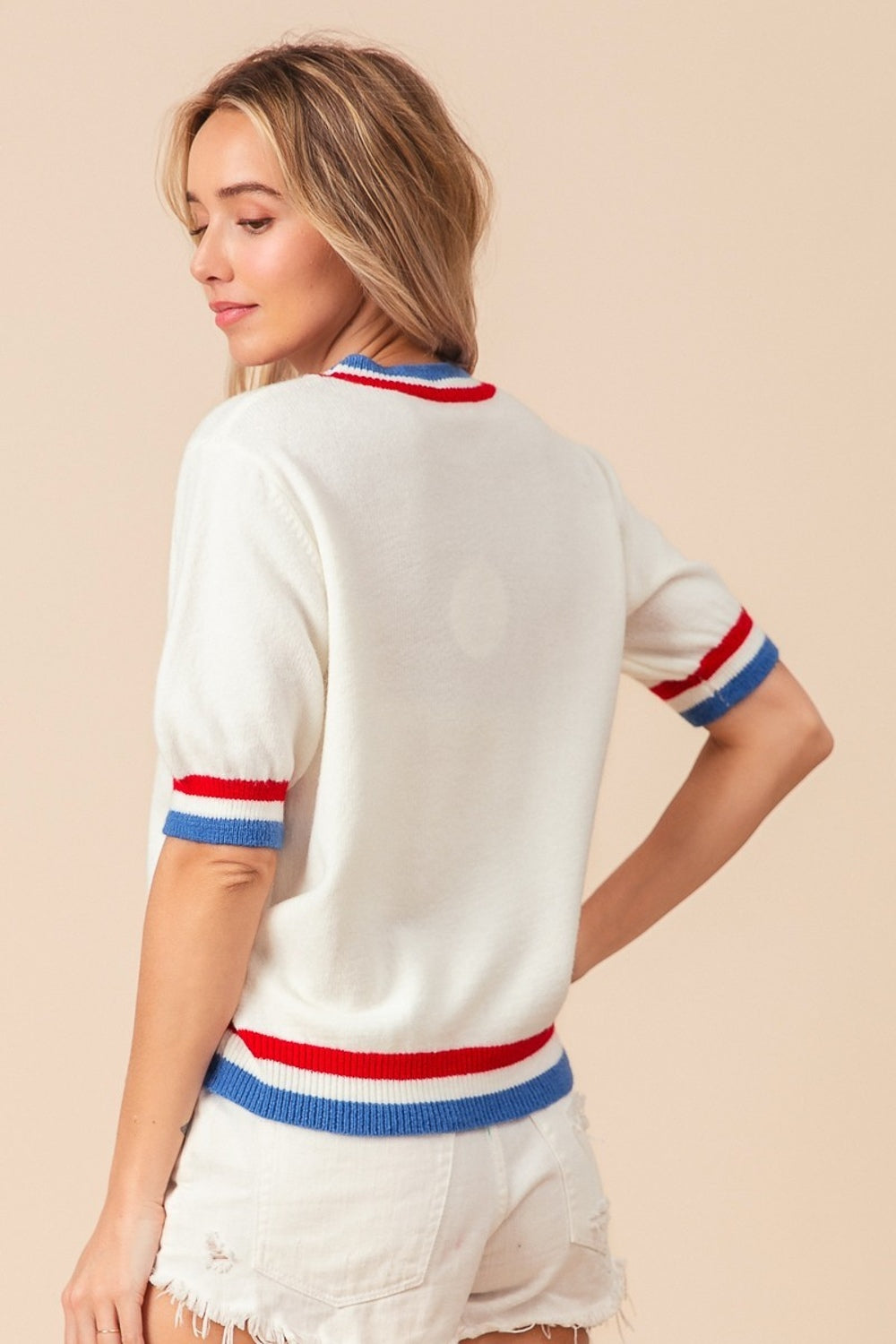 A person wearing the BiBi US Flag Theme Striped Heart Sweater, featuring a white short-sleeve design with red and blue stripes resembling the US flag and adorned with a red heart motif. They are also wearing white shorts and smiling with hands on hips.