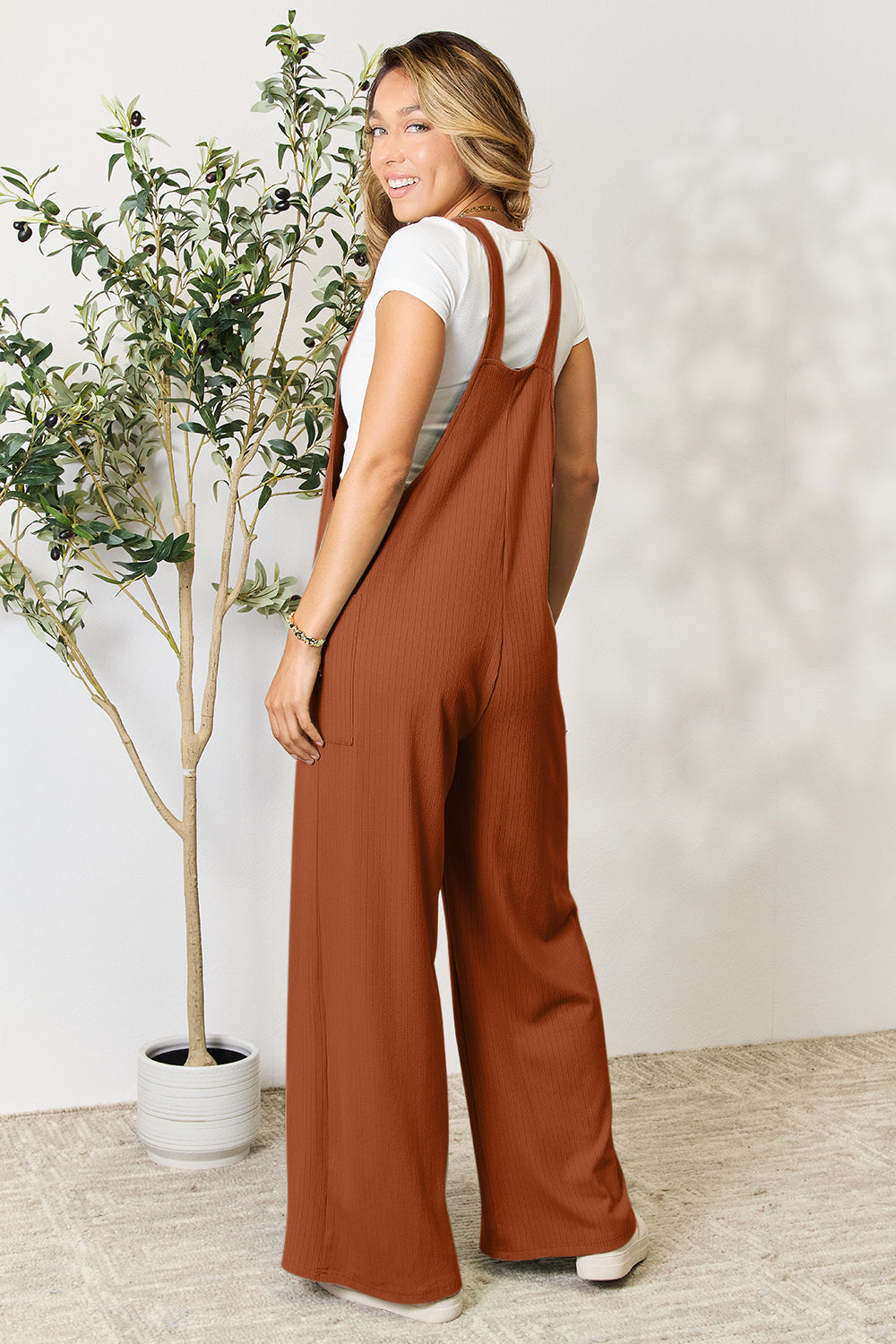 A smiling person wearing the "Double Take Full Size Wide Strap Overall with Pockets," known for its basic style and size-inclusive fit, stands indoors next to a potted plant.