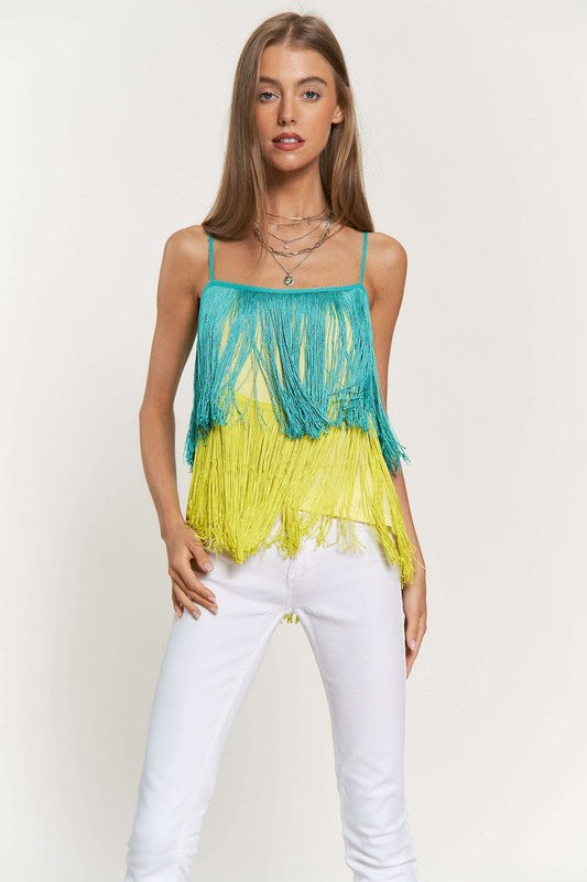 A woman with long brown hair is wearing a Fringe Overlay Cross Straps Party Cami Top featuring blue and yellow layers, paired with white pants, standing against a plain background.