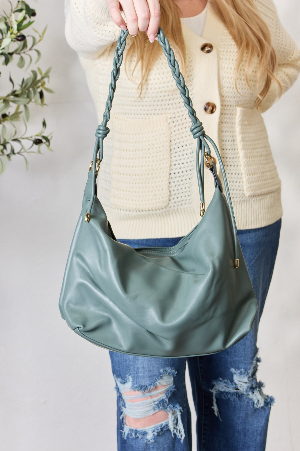 A person in a knitted sweater and jeans holds the SHOMICO Braided Strap Shoulder Bag, a blue-green statement accessory featuring straps crafted from vegan leather.