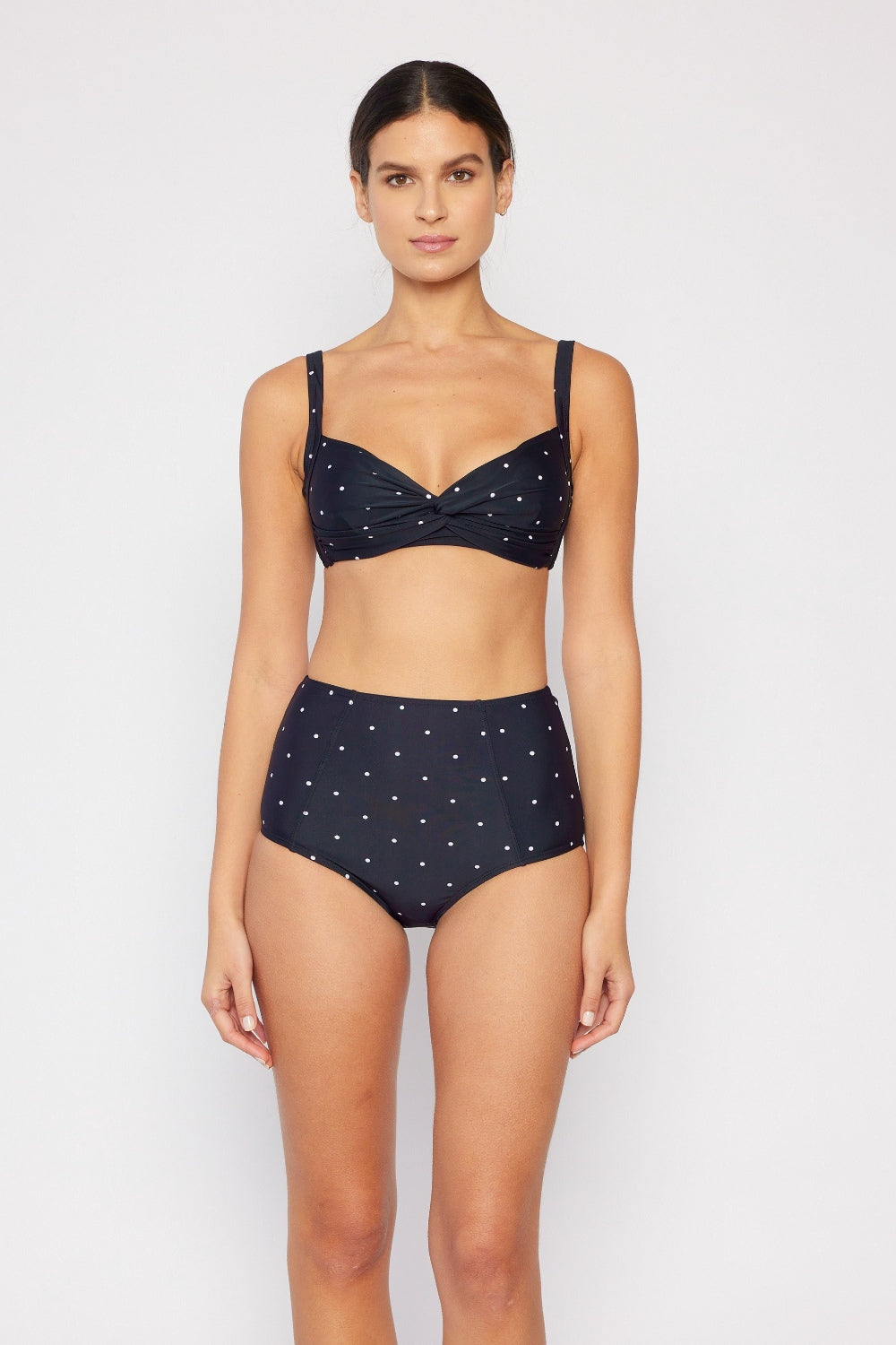 Wearing the fashionable Marina West Swim Take A Dip Twisted High-Rise Bikini Set in black polka dot, one effortlessly combines style with practicality. This chic bikini is complemented by a woven bag with striped handles, set against a neutral backdrop for the ultimate summer vibe.