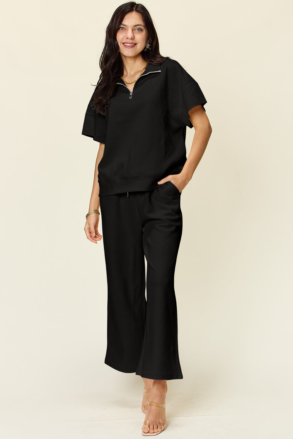 A woman is wearing the Double Take Full Size Texture Half Zip Short Sleeve Top and Pants Set, featuring a white zip-up short-sleeve top and matching wide-leg, drawstring pants. She stands with one hand in her pocket and smiles against a light background.