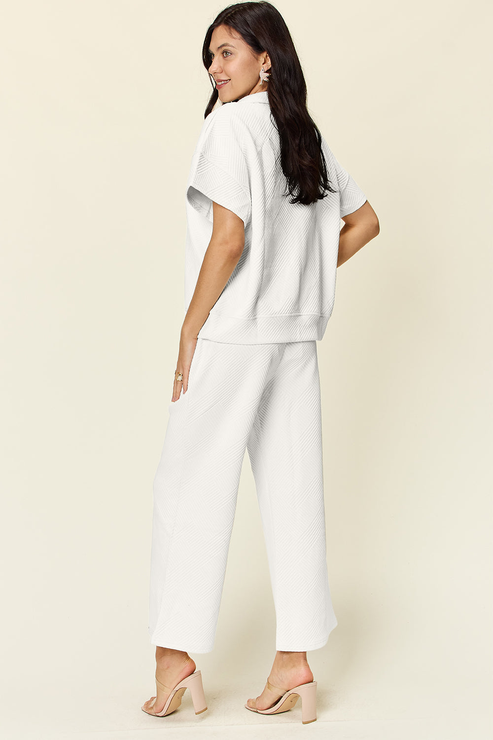 A woman is wearing the Double Take Full Size Texture Half Zip Short Sleeve Top and Pants Set, featuring a white zip-up short-sleeve top and matching wide-leg, drawstring pants. She stands with one hand in her pocket and smiles against a light background.