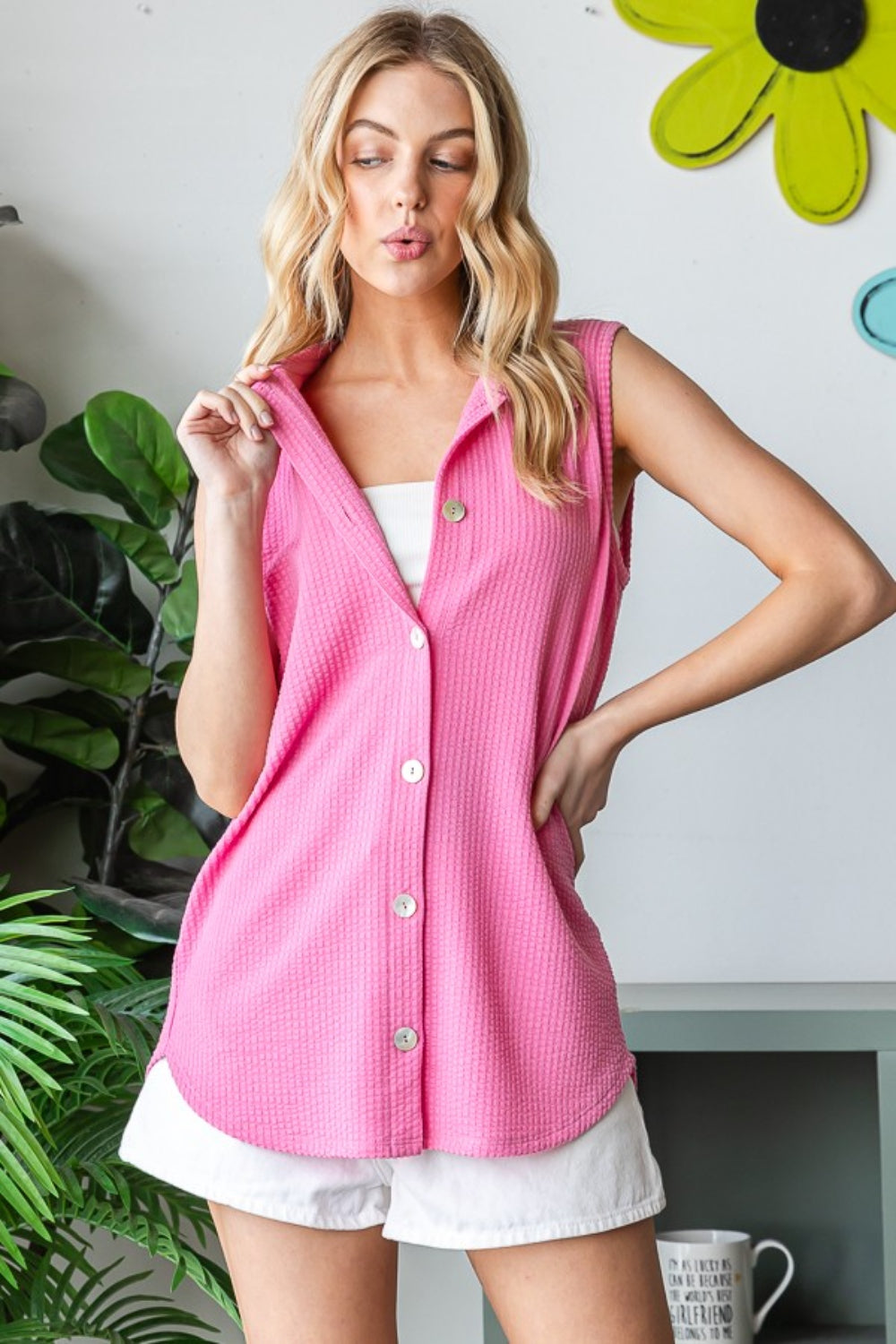 A woman models the Heimish Full Size Texture Button Up Sleeveless Top in pink paired with white shorts, posing indoors amidst green plants and vibrant yellow flower wall decor, highlighting a versatile style.