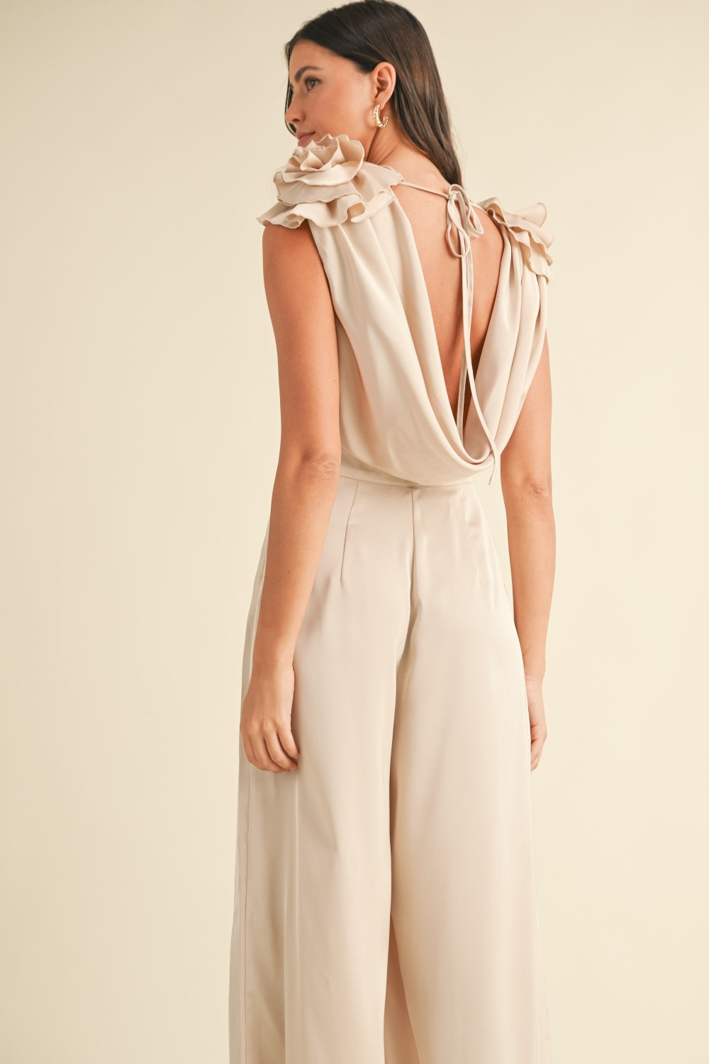 A woman stands wearing the MABLE 3D Floral Applique Deep Cowl Neck Jumpsuit, which showcases a light beige color with a draped neckline and ruffled sleeves. Featuring a wide leg silhouette, the outfit flows elegantly. She has long hair and is wearing high heels.