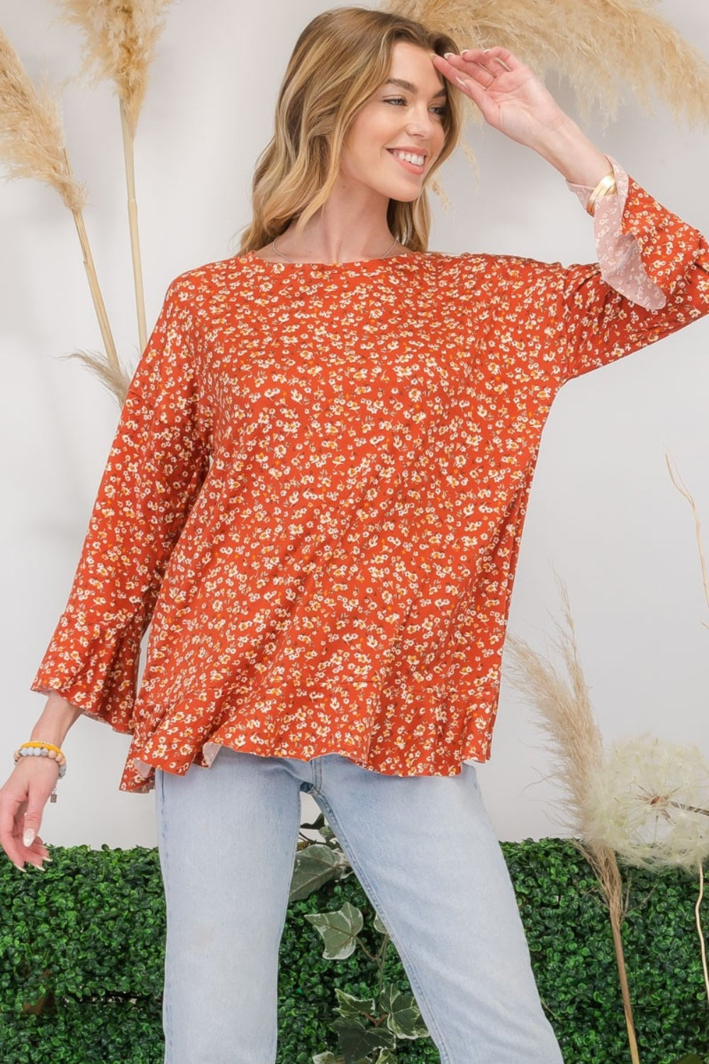 A woman wearing the Celeste Full Size Floral Ruffle Detail Top and jeans stands smiling in front of pampas grass and greenery, exuding feminine flair.