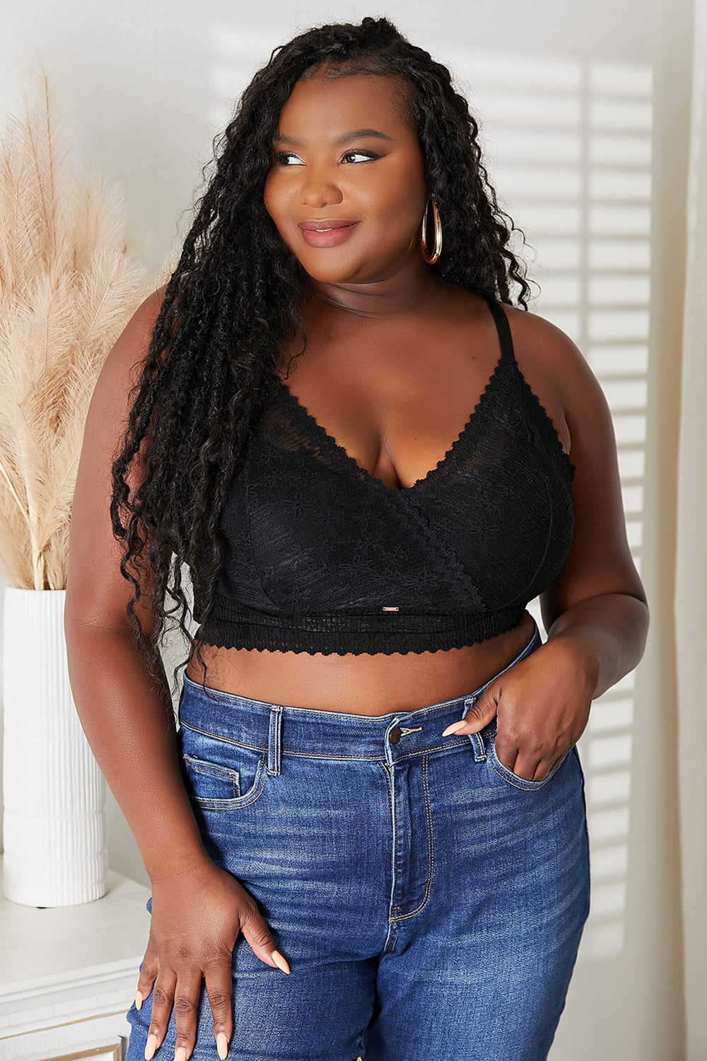 A woman with long black braids is smiling while wearing the JadyK Skye Full Size Lace Bralette and blue jeans, standing near a plant in a white pot.