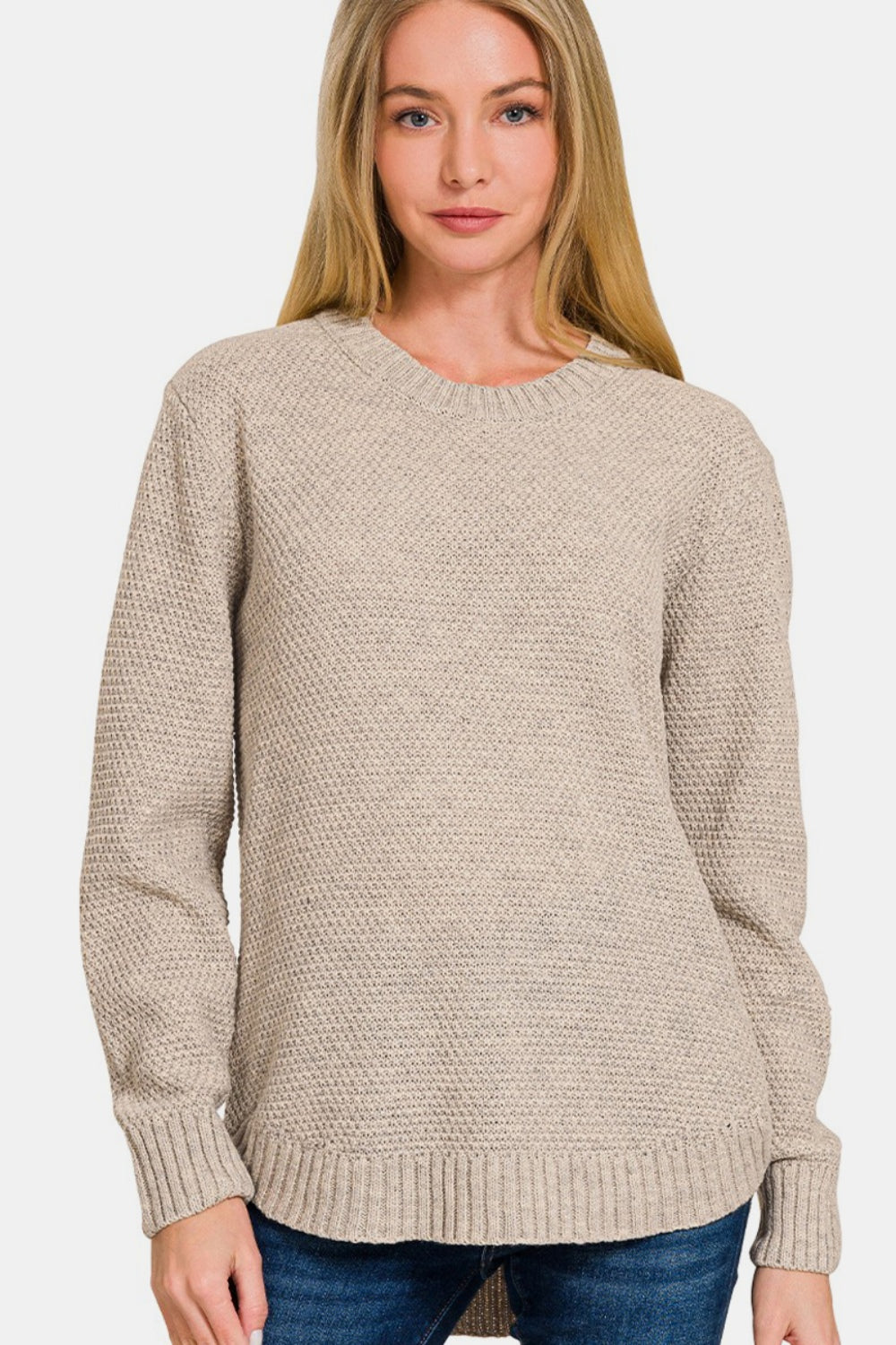 A person wearing a Zenana Full Size Round Neck Long Sleeve Curved Hem Sweater and jeans smiles while gently holding their neck.