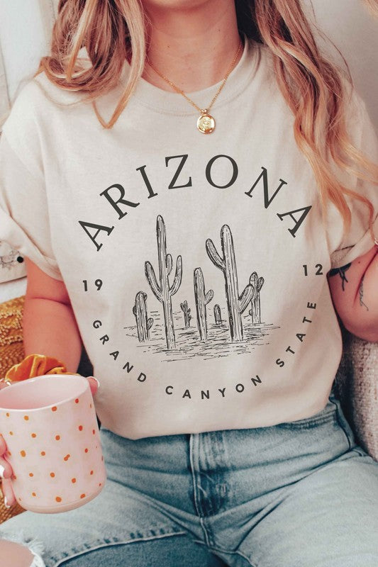 Person wearing an ARIZONA GRAND CANYON STATE Graphic Tee, made of premium 100% cotton with a design featuring the words "ARIZONA GRAND CANYON STATE 1912" and a drawing of cacti, paired with white shorts.