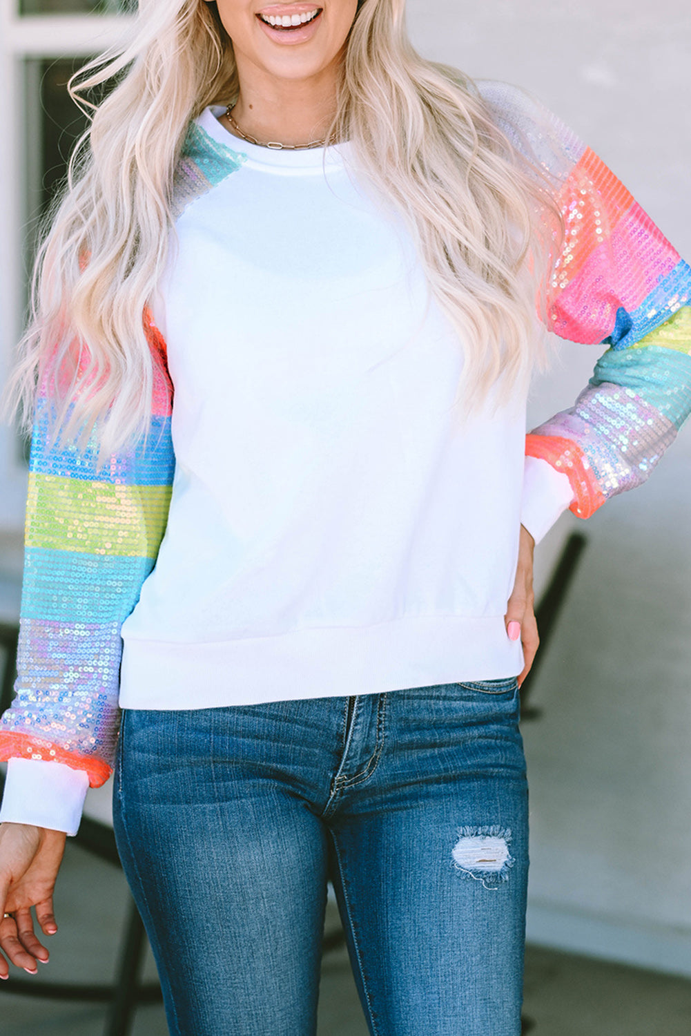 A person with long blonde hair is seen from the back, wearing a White Sequin Color Block Raglan Sleeve Pullover Sweatshirt and blue jeans. They are standing in front of a building with large windows.