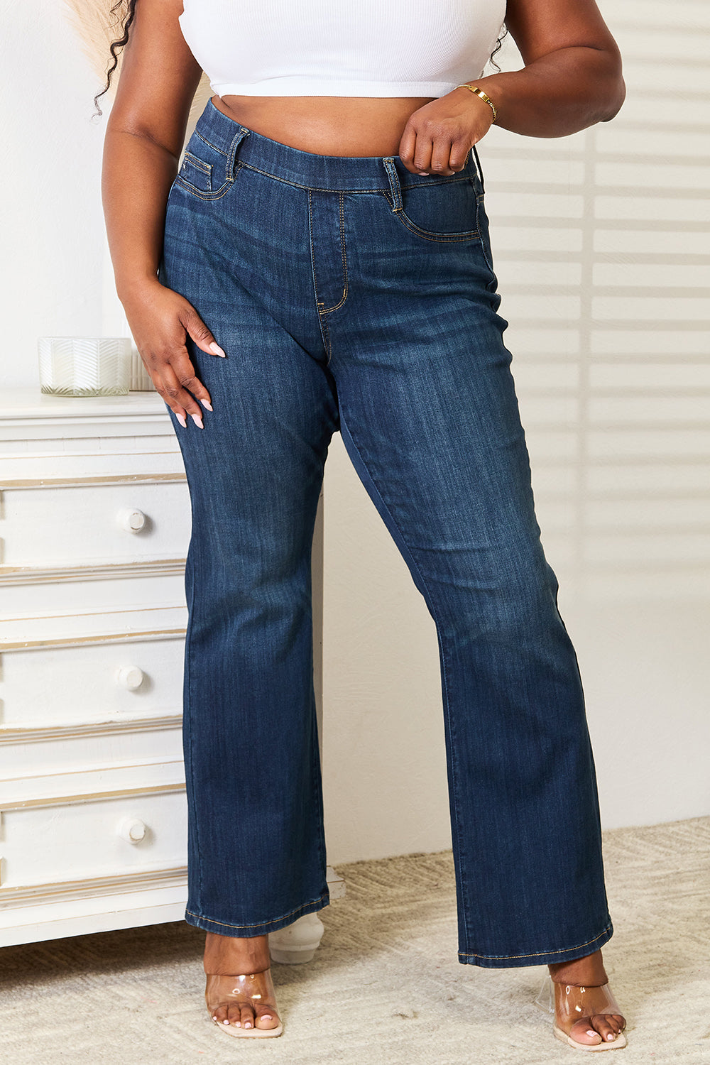 A person wearing a white crop top and Judy Blue Full Size Elastic Waistband Slim Bootcut Jeans stands next to a white dresser.