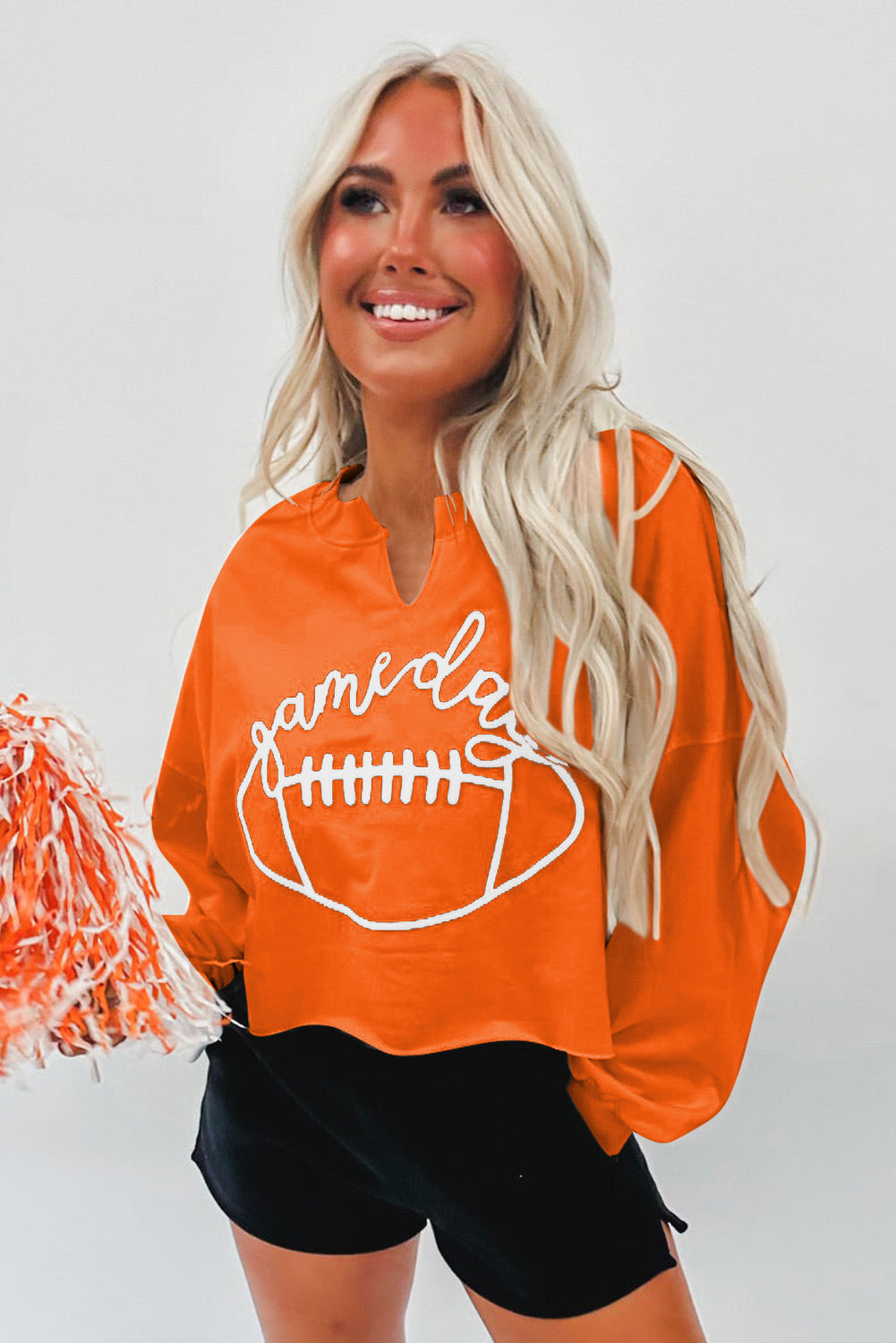 A woman with blonde hair, wearing an Orange Game Day Lettering Rugby Football Notched Neck Sweatshirt and blue jeans, stands next to a decorated Christmas tree.