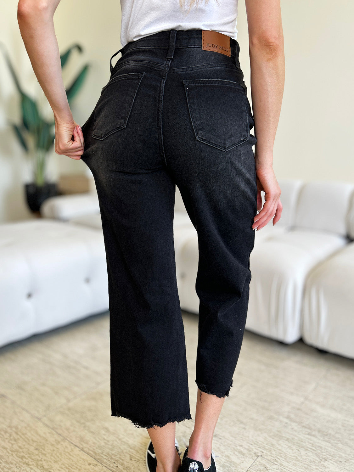 Person wearing Judy Blue Full Size High Waist Button Fly Jeans and black sneakers, standing indoors. Hands are in pockets, and the background includes a white couch and a plant. These high-waisted jeans make the outfit sleek and stylish.