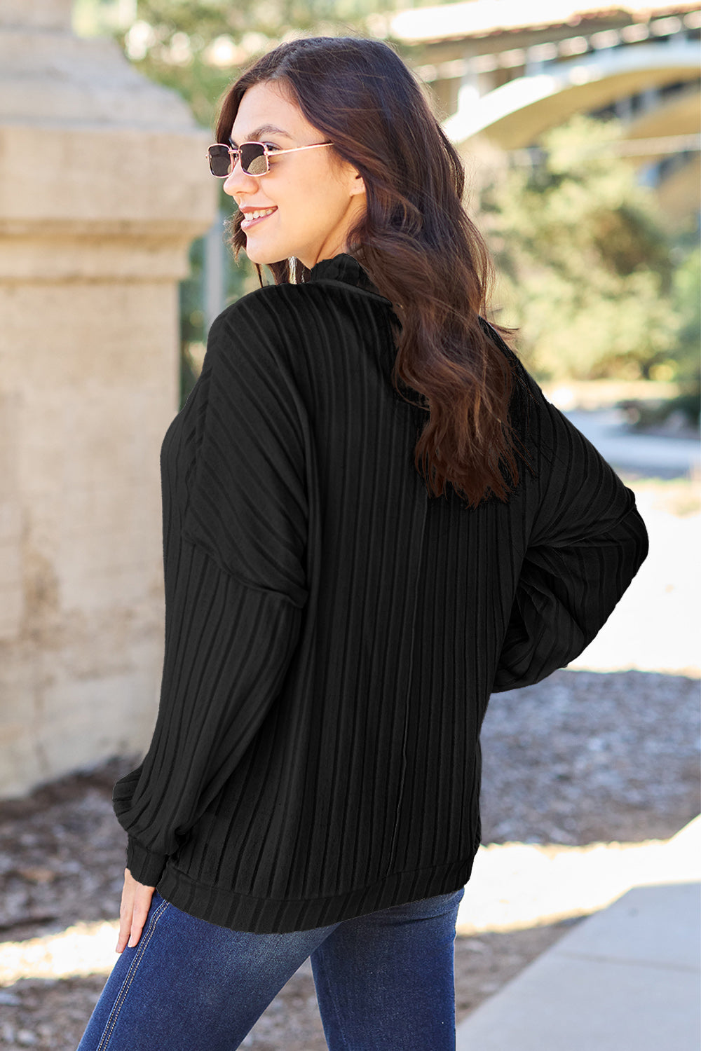 A woman with long hair is wearing a green Basic Bae Full Size Ribbed Exposed Seam Mock Neck Knit Top, blue jeans, and sunglasses. She is standing outdoors and smiling.