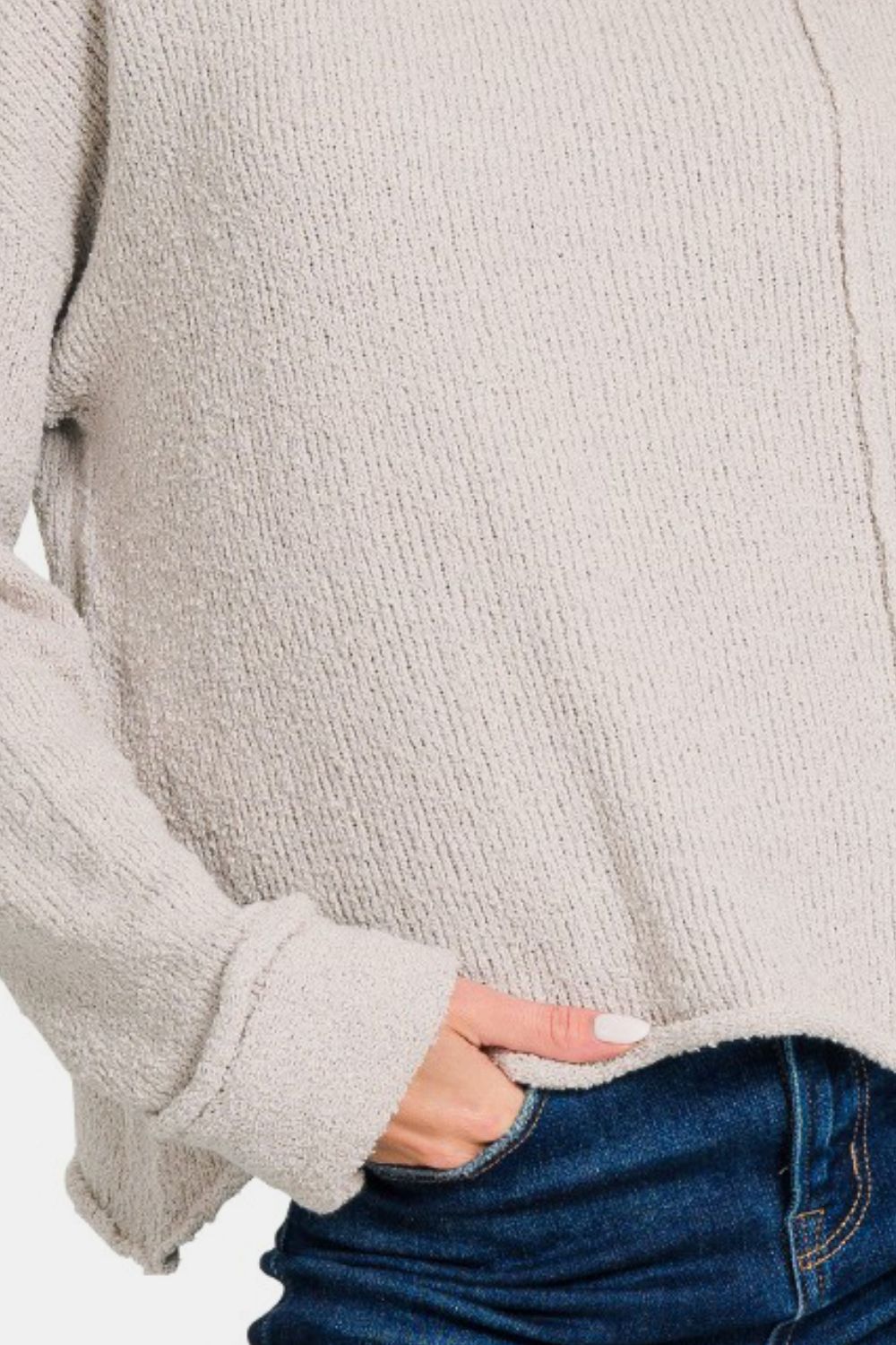 A woman in a light beige Zenana Asymmetric Hem Drop Shoulder Sweater poses with a hand near her head and another in her pocket, smiling slightly. She is wearing blue jeans against a plain white background.