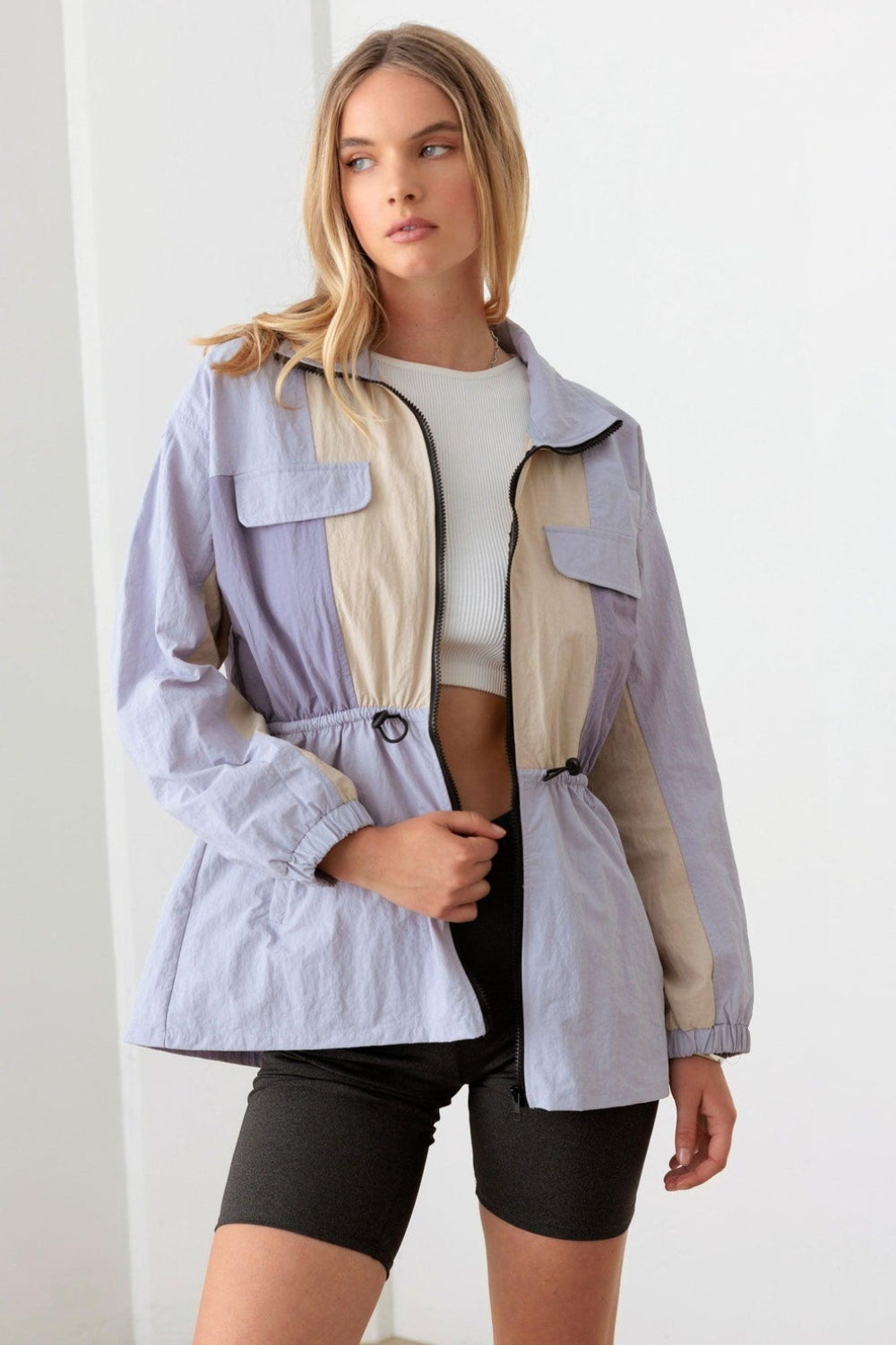 A woman wearing the Le Lis Color Block Collared Wind Breaker Jacket over a white crop top and black shorts stands in a well-lit room.