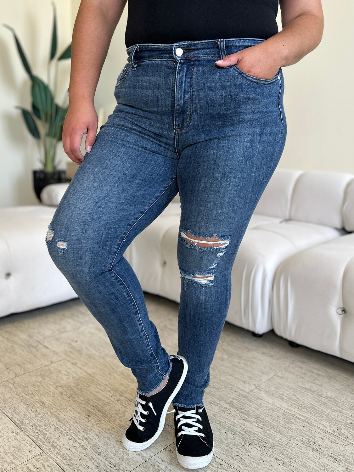 A person wearing Judy Blue Full Size High Waist Distressed Skinny Jeans and black sneakers stands indoors on a wooden floor, with a white sofa and green plant in the background.